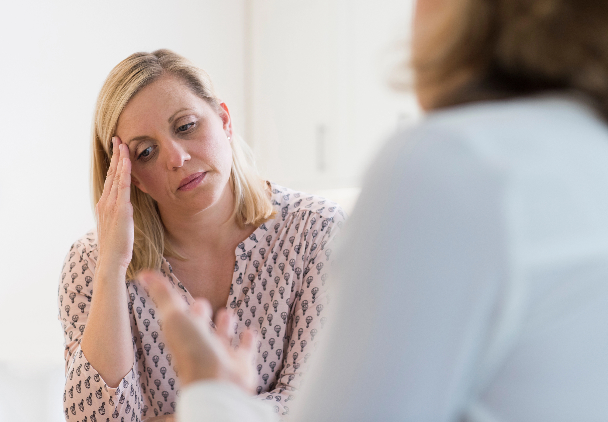 A woman with blonde hair rests her hand on her forehead, appearing stressed or concerned, while listening to another person in the foreground who is gesturing with their hands. The setting seems to be an indoor environment, possibly an office or home.