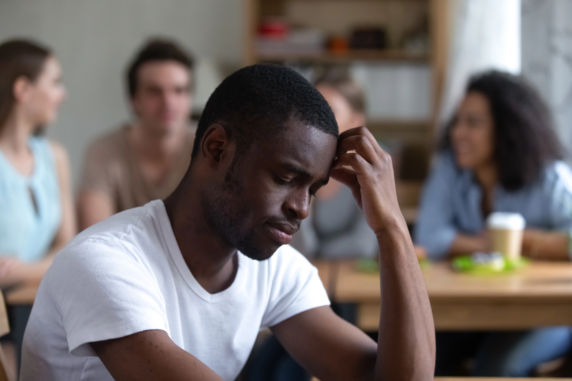 A young man in a white t-shirt sits alone with a thoughtful expression in a busy room. In the background, a group of three people are engaged in conversation, slightly out of focus. The room has a casual atmosphere with bookshelves and tables.