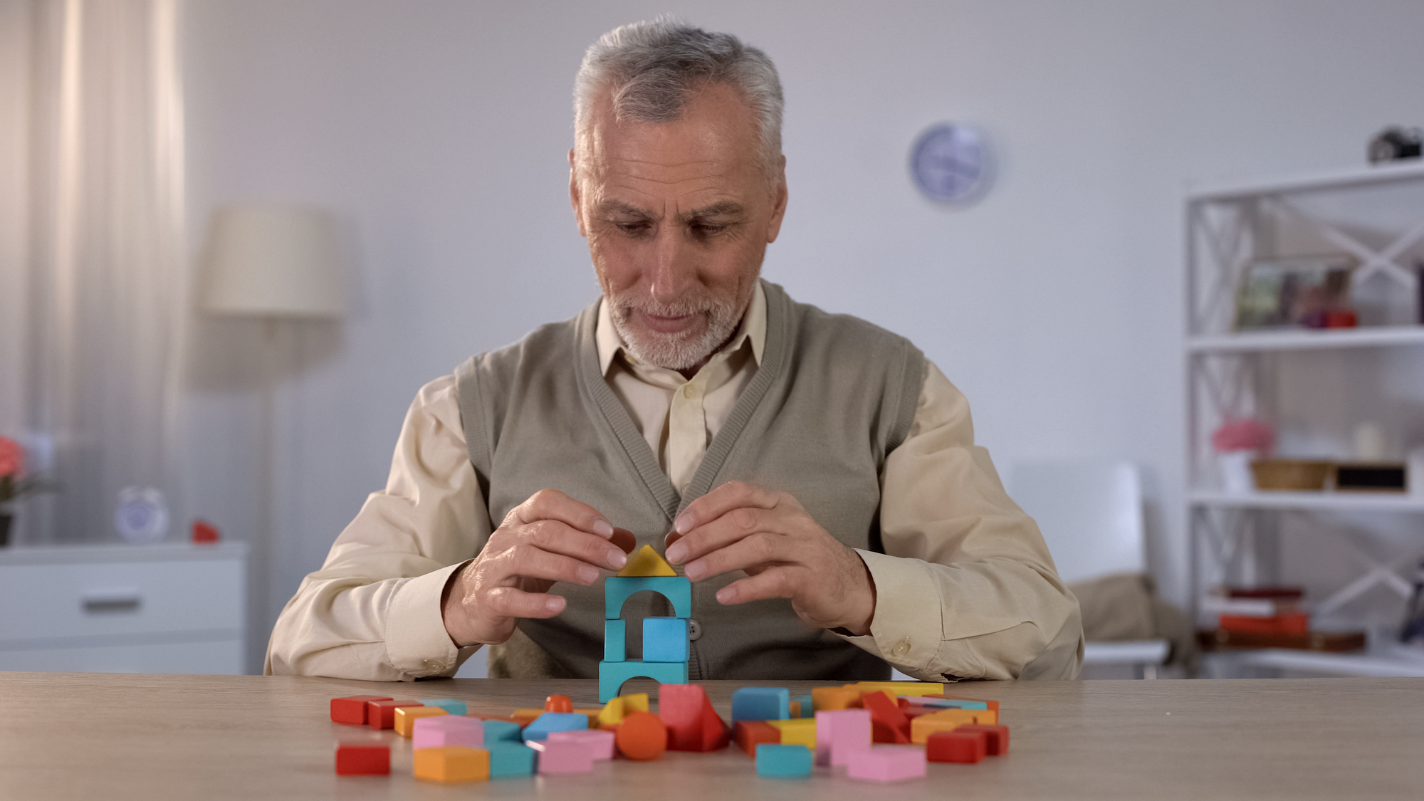 An elderly man with gray hair and a beard is seated at a table, carefully building a structure with colorful wooden blocks. He is wearing a beige sweater vest over a light-colored shirt. The room is softly lit, with a lamp and shelves in the background.