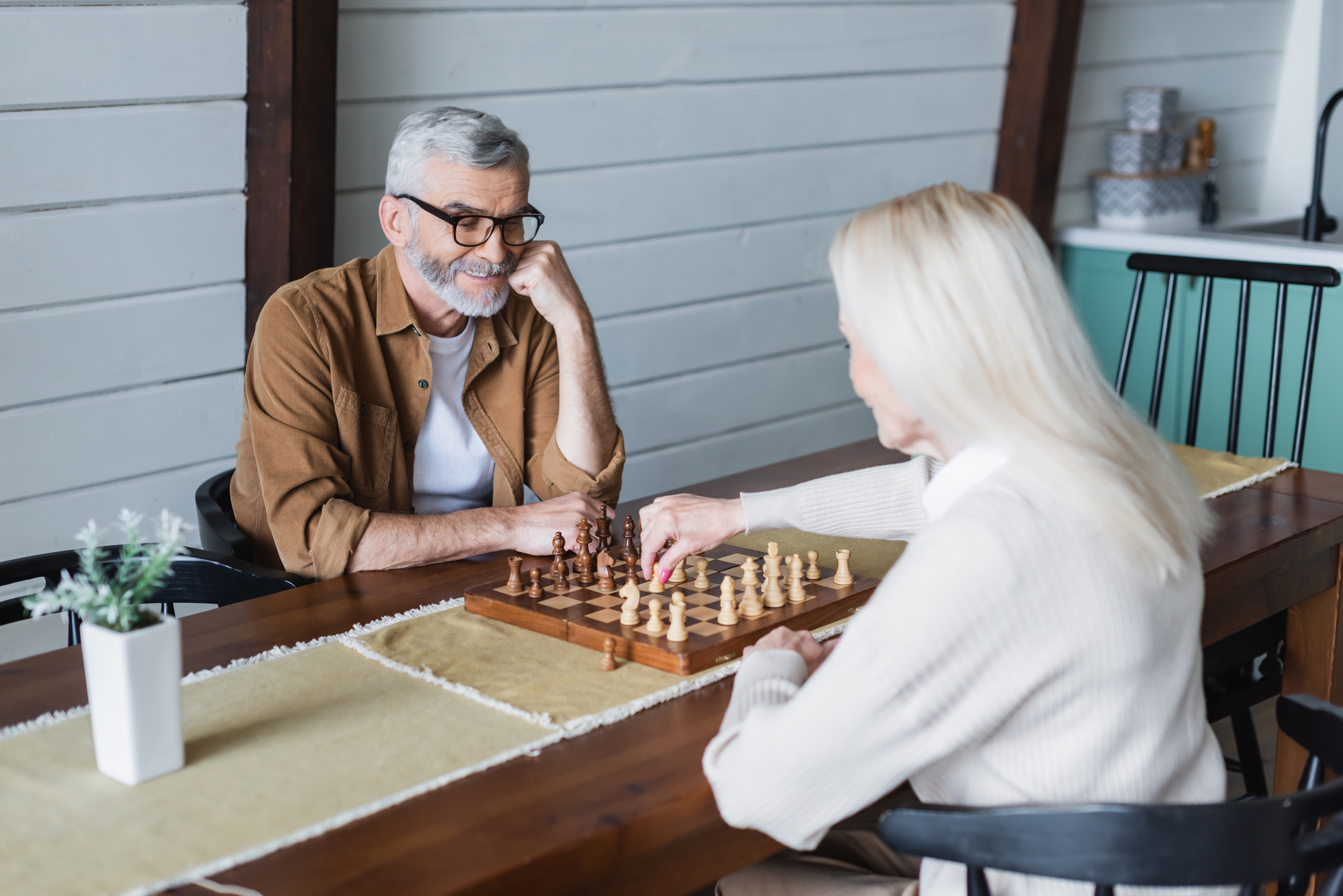 An elderly couple is engaged in a chess game at a wooden table inside a cozy, well-lit room. The man with gray hair and a beard looks at the board thoughtfully, while the woman with long blonde hair makes a move. A potted plant decorates the table.