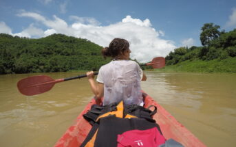 A person with wet hair and a white shirt is seen kayaking on a calm river, surrounded by lush green hills. They are paddling with red oars under a partly cloudy blue sky. Brightly colored life jackets are on the kayak’s deck.