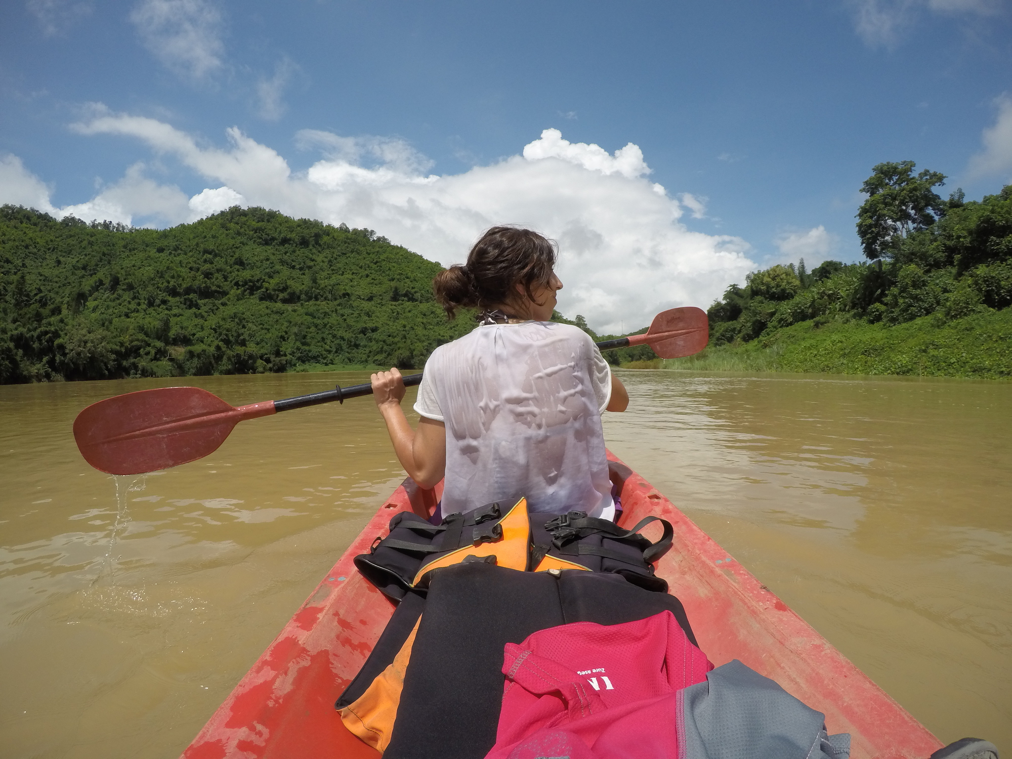 A person with wet hair and a white shirt is seen kayaking on a calm river, surrounded by lush green hills. They are paddling with red oars under a partly cloudy blue sky. Brightly colored life jackets are on the kayak’s deck.