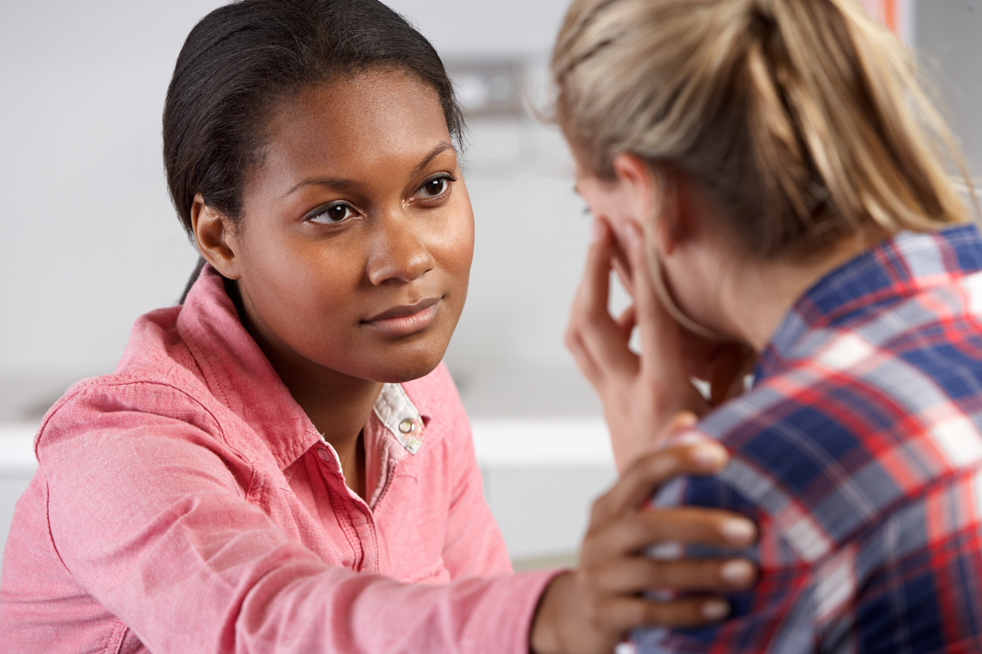 A woman in a pink shirt is offering comfort by gently placing her hand on the shoulder of another woman who is wiping her eyes with a tissue and appears upset. They are indoors, and the background is slightly blurred.