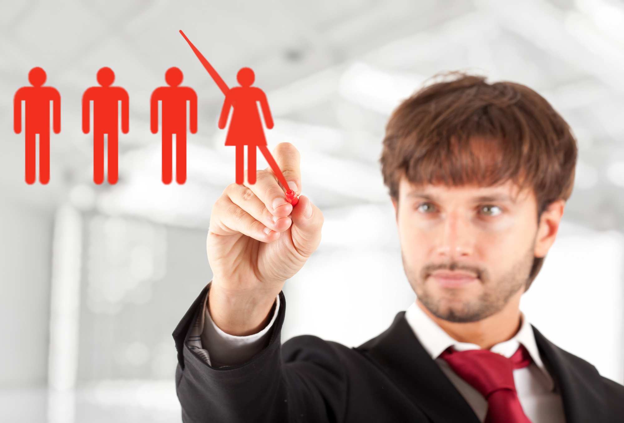 A man in a suit with a red tie is using a red marker to point at a transparent screen featuring four red human icons, with one being female and the rest male. The background is a blurred office setting with bright natural light.