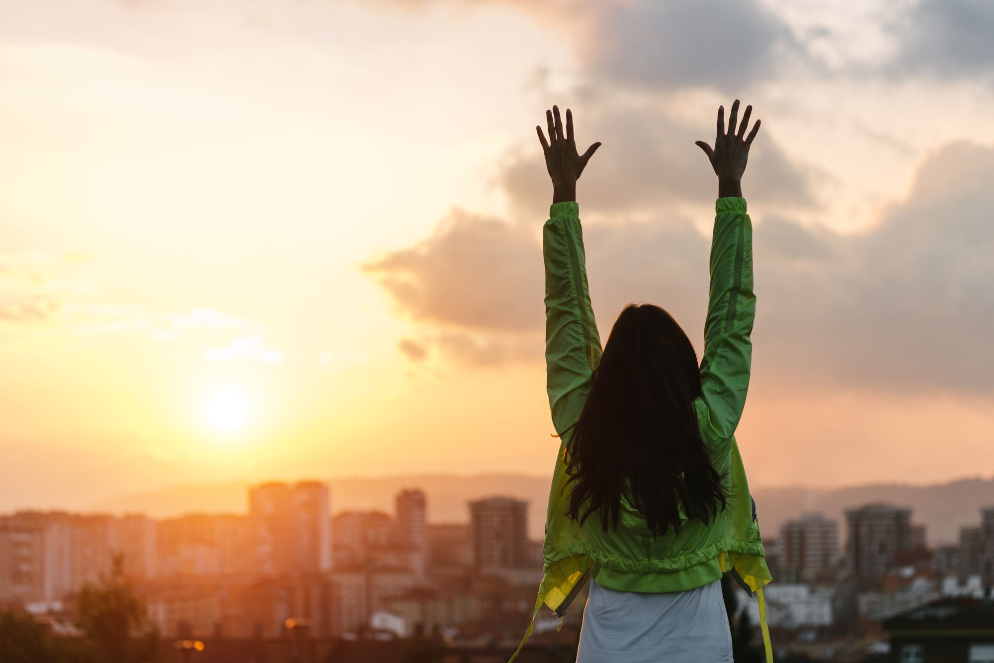 A person with long hair, wearing a green jacket, stands with their back to the camera, arms raised towards the sky at sunset. The city skyline and buildings are silhouetted against the colorful sky in the background.