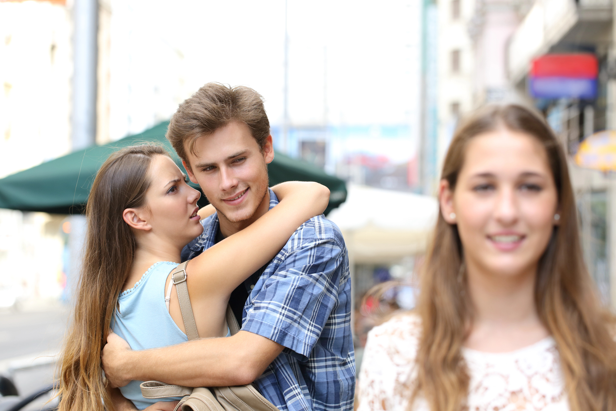 A woman in focus looks ahead and smiles, while in the background, a couple shares an embrace with the woman looking at the man. The scene is set outdoors on a street with buildings and an outdoor umbrella.