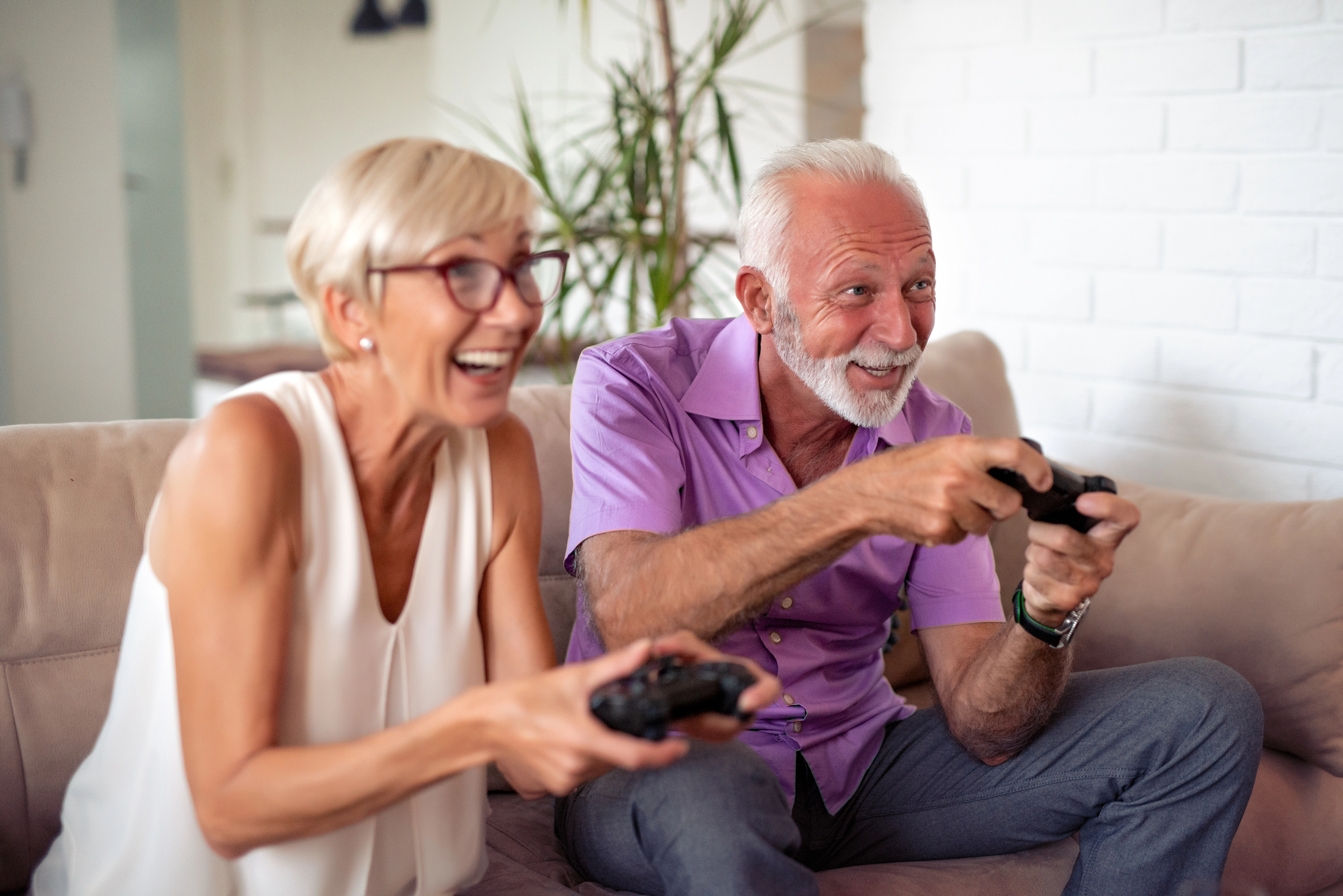 An elderly couple sits on a couch, both smiling and holding video game controllers. The woman on the left has short blonde hair and glasses, wearing a white blouse. The man on the right has white hair and a beard, wearing a purple shirt. A plant is in the background.