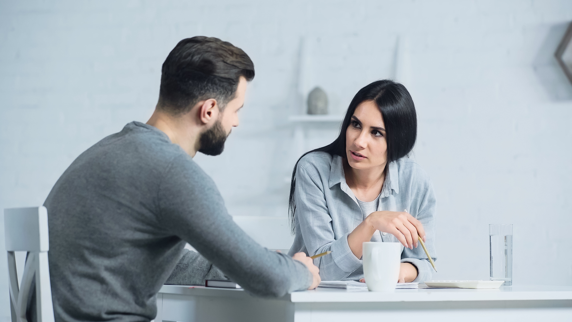 A man and a woman are engaged in a serious conversation at a white table in a minimalist room. Both are holding pens and have papers in front of them. The woman is gesturing with her pen, while the man listens attentively. There are white mugs on the table.