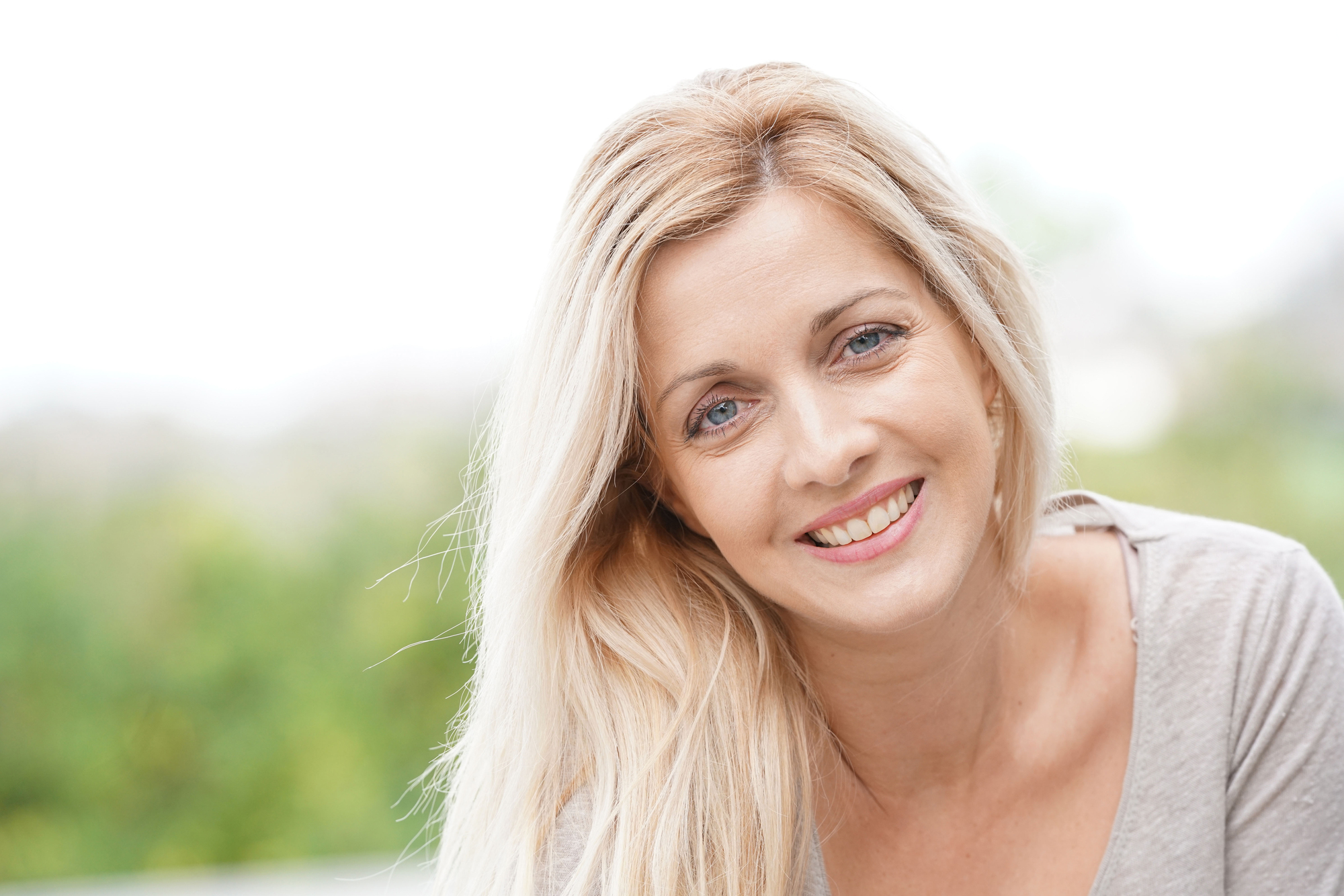 A woman with long blonde hair and blue eyes is smiling while looking at the camera. She is wearing a light gray top. The background is blurred with greenery, suggesting an outdoor setting.