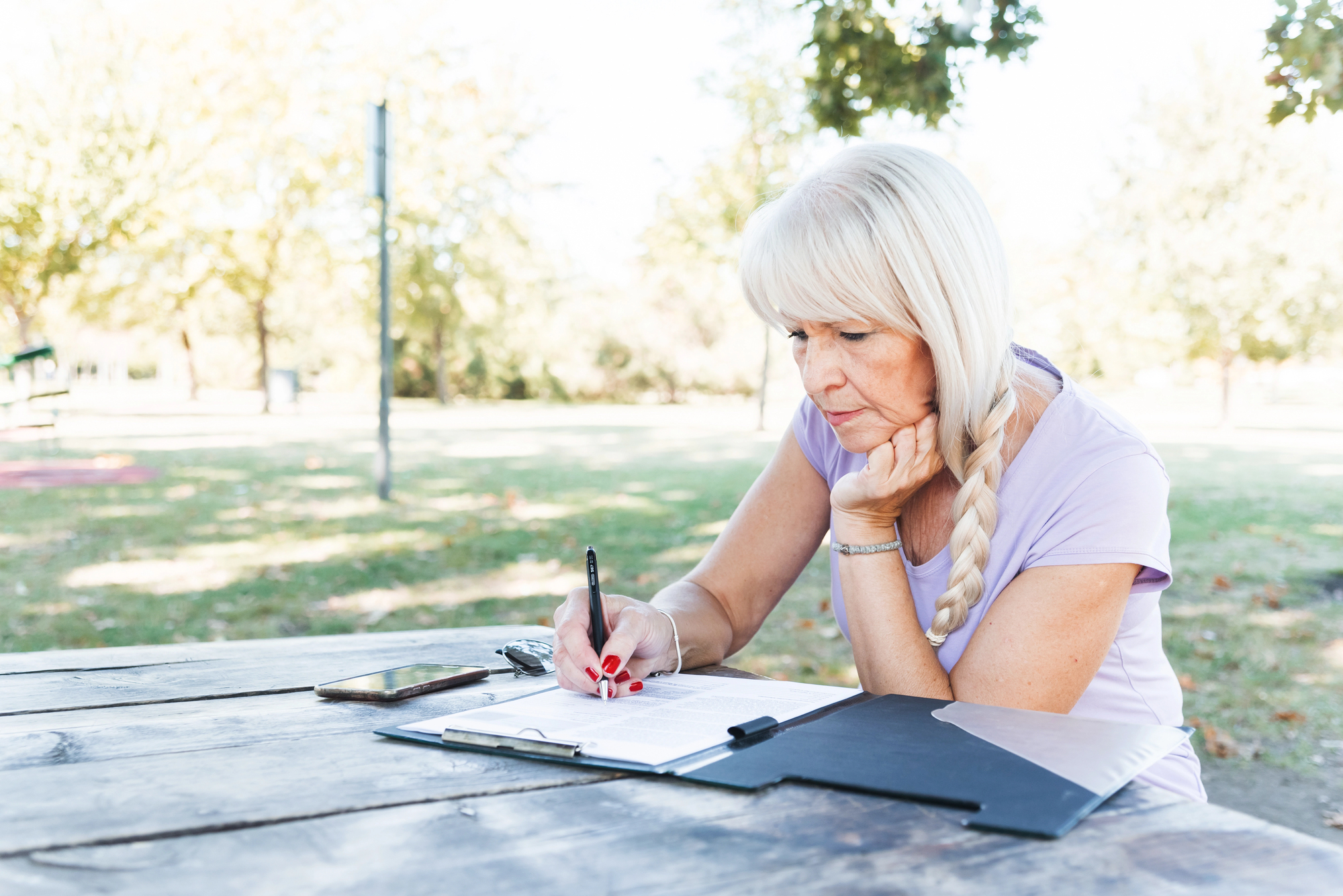 A older woman with long, blonde hair sits at a wooden table outdoors, writing on documents in a folder. She is wearing a light purple shirt and has a smartphone placed on the table next to her. Trees and a grassy area appear in the background.