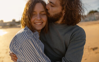 A couple embraces on a sunny beach. The woman with short hair smiles with her eyes closed, wearing a striped shirt. The man with a beard kisses her on the cheek, wearing a gray sweatshirt. The background shows sandy beach and blurred cityscape.