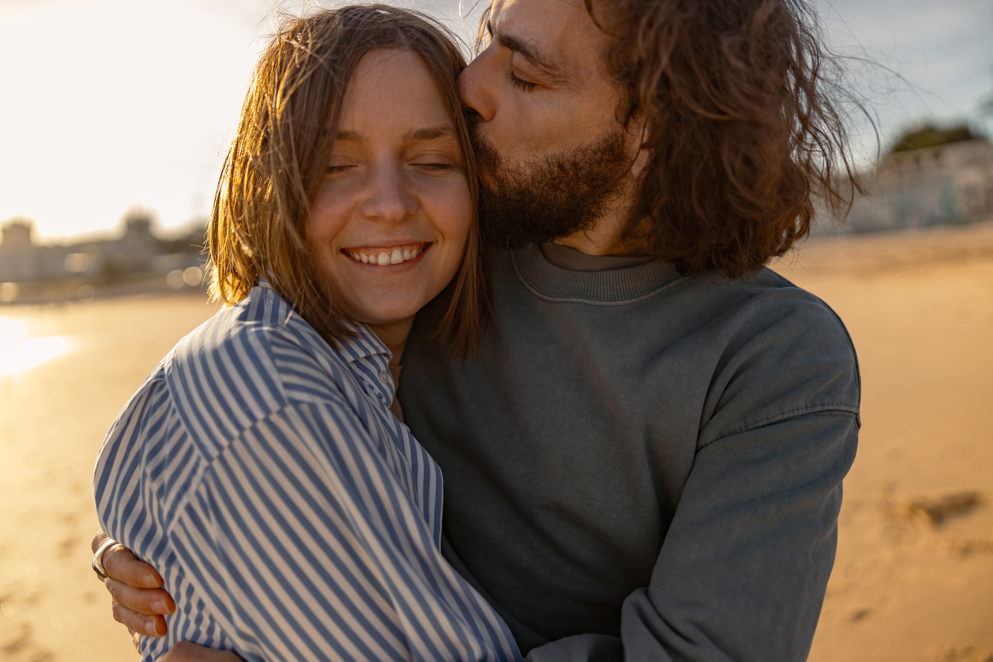 A couple embraces on a sunny beach. The woman with short hair smiles with her eyes closed, wearing a striped shirt. The man with a beard kisses her on the cheek, wearing a gray sweatshirt. The background shows sandy beach and blurred cityscape.