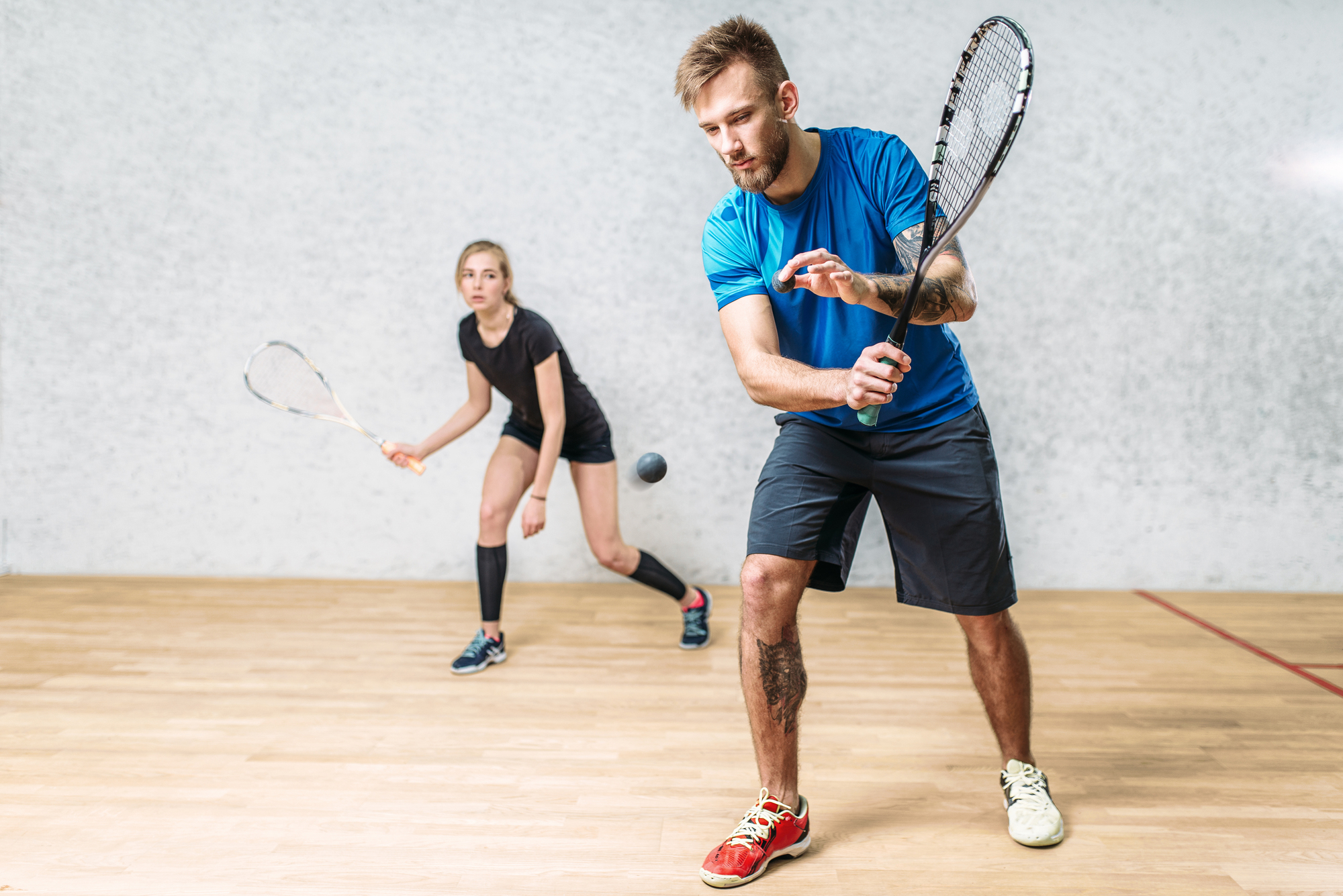 Two squash players are in action on the court. The male player in the foreground, wearing a blue shirt and black shorts, is poised to hit the ball with his racket. The female player in the background, dressed in black, is preparing for the next move.