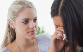 A blonde woman with a concerned expression comforts a brunette woman who is crying and wiping her tears with a tissue. The scene appears to show support and empathy between the two women.