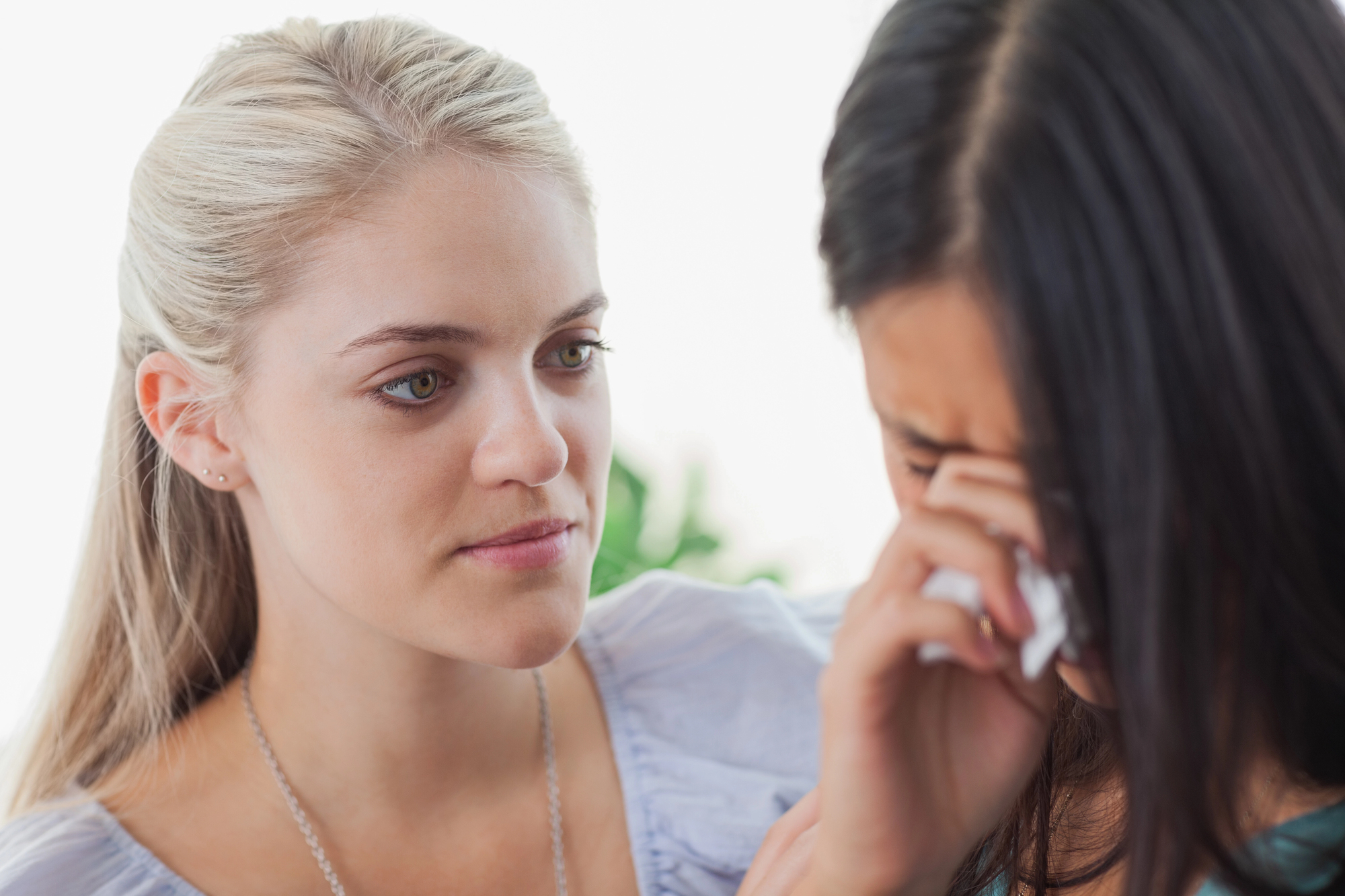 A blonde woman with a concerned expression comforts a brunette woman who is crying and wiping her tears with a tissue. The scene appears to show support and empathy between the two women.