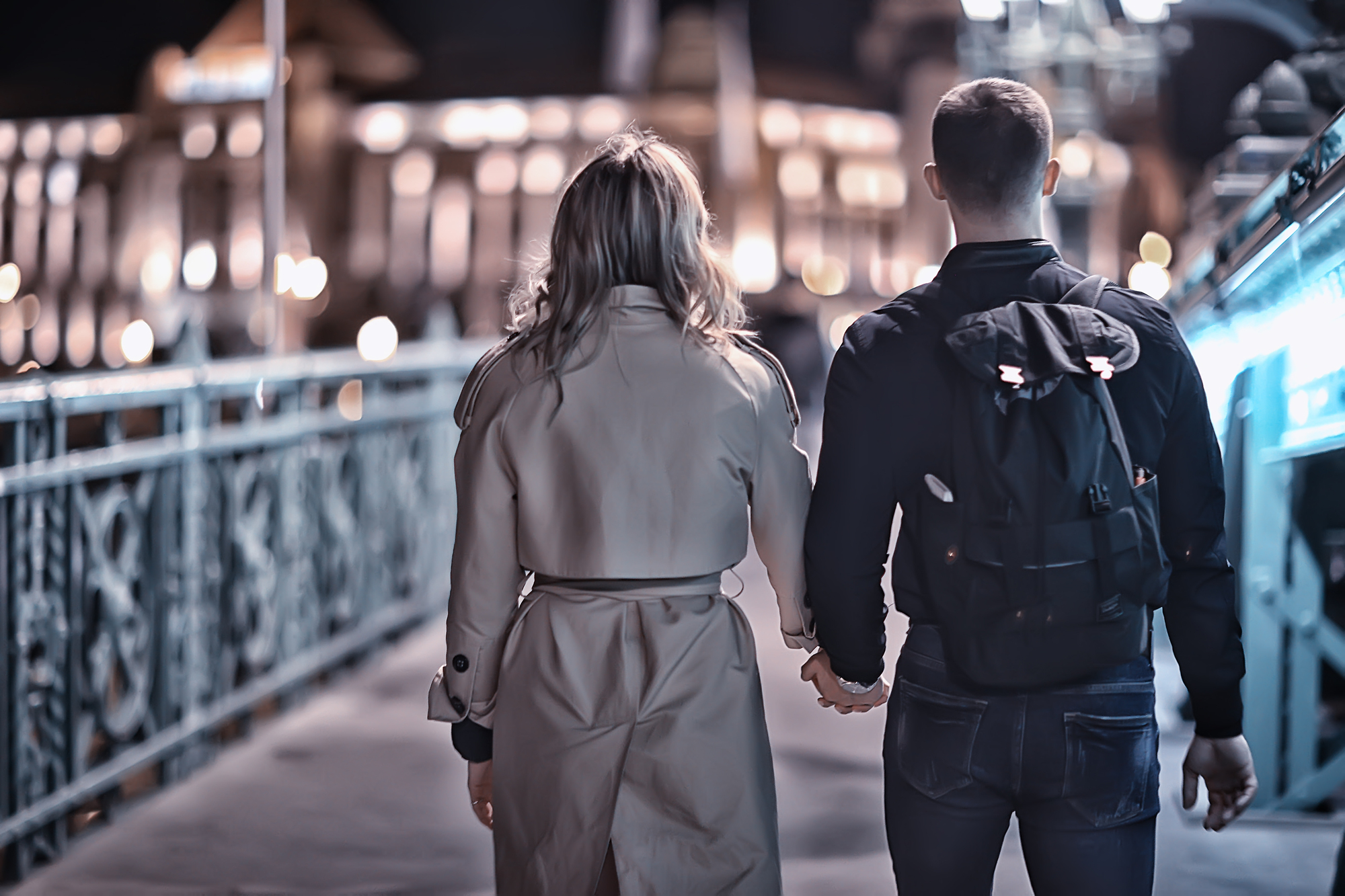 A couple walks hand-in-hand on a bridge at night. The woman wears a light-colored trench coat, while the man wears dark clothing and carries a backpack. The bridge is adorned with ornate railings and illuminated by soft lighting. A blurred cityscape is visible in the background.