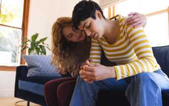 Two people sit closely on a couch, with one person, wearing a yellow and white striped shirt, looking down and appearing distressed. The other person, with long hair and a beige sweater, has an arm around their shoulder, offering comfort. A plant is visible in the background.