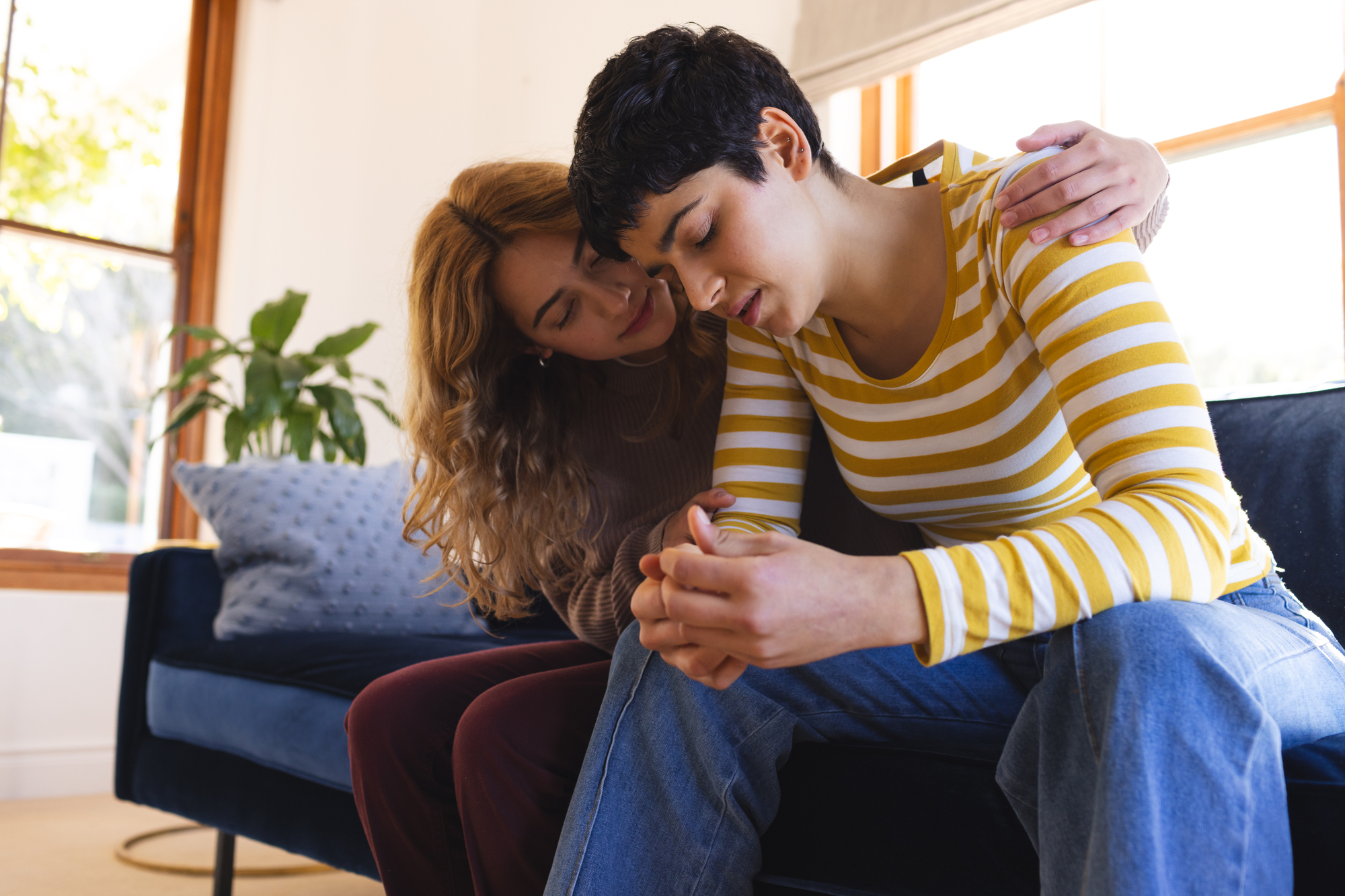 Two people sit closely on a couch, with one person, wearing a yellow and white striped shirt, looking down and appearing distressed. The other person, with long hair and a beige sweater, has an arm around their shoulder, offering comfort. A plant is visible in the background.