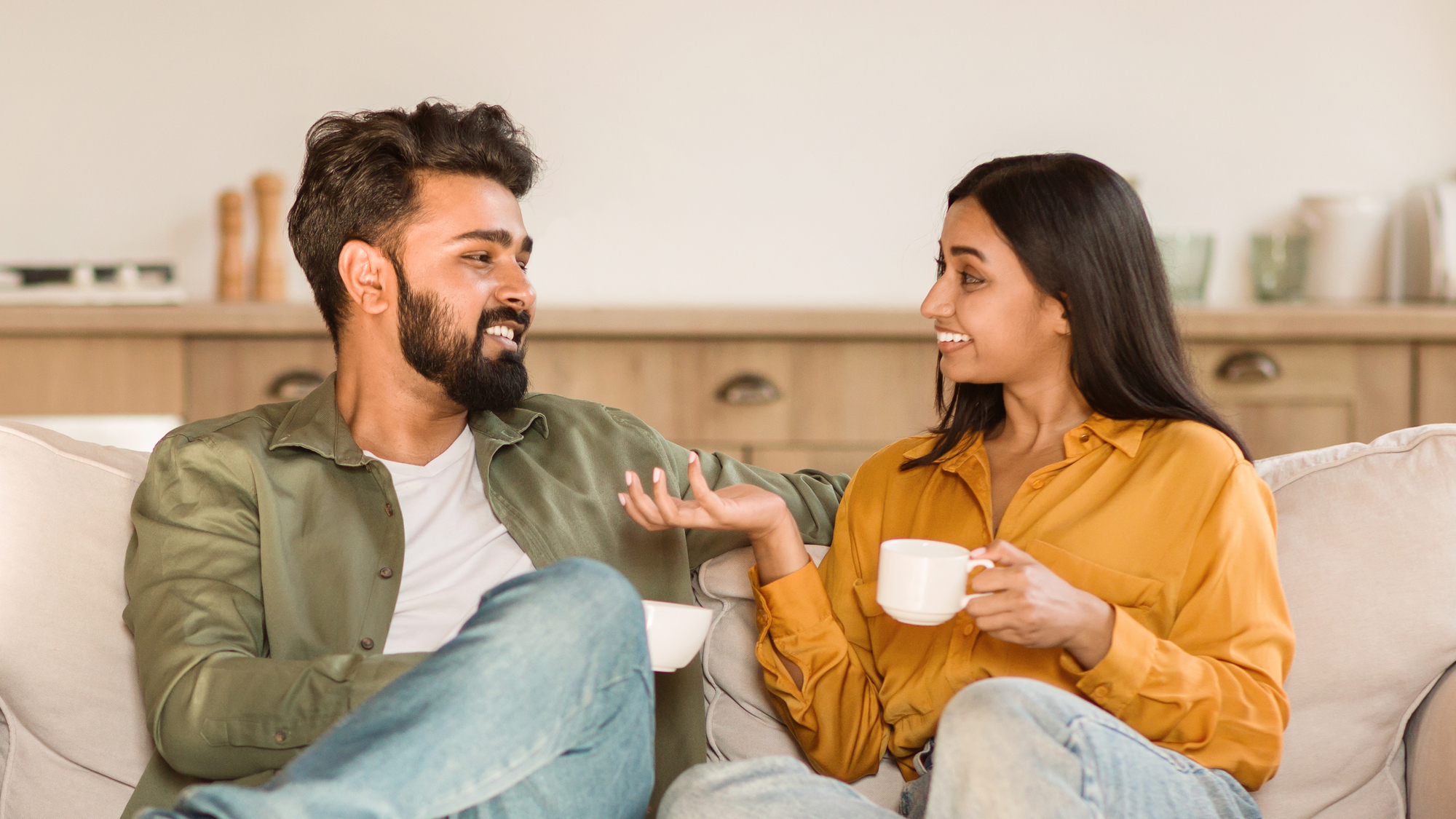 A man and woman are sitting on a couch, facing each other, and smiling. The man has a beard and is wearing a green shirt, while the woman has long dark hair and is wearing a mustard-colored blouse. They both are holding white mugs, possibly enjoying a conversation over coffee or tea.