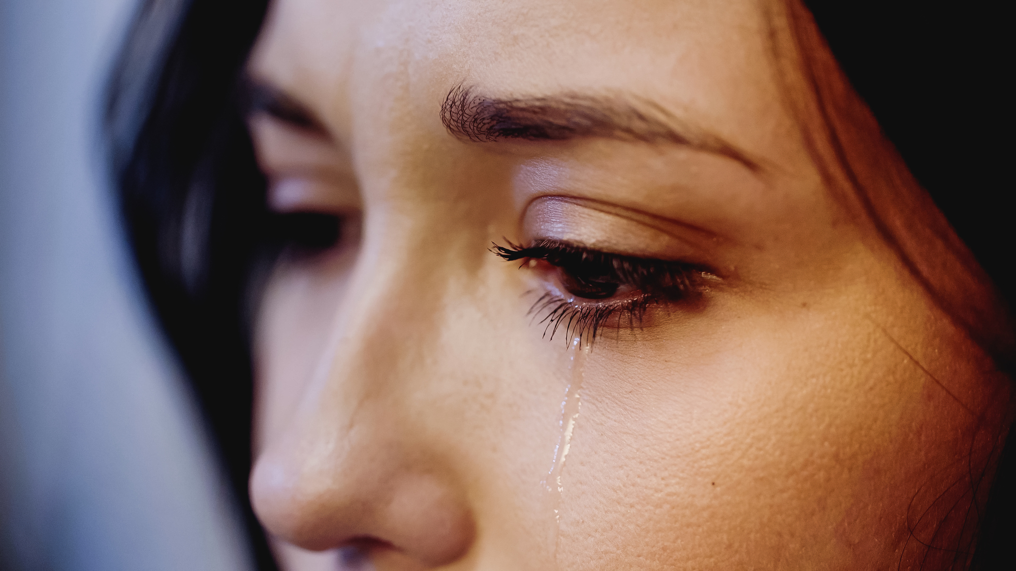 Close-up of a person's face showing an eye and eyebrow, with a single tear running down their cheek. The person has dark hair and appears to be in a moment of sadness or emotional reflection. The background is blurred.