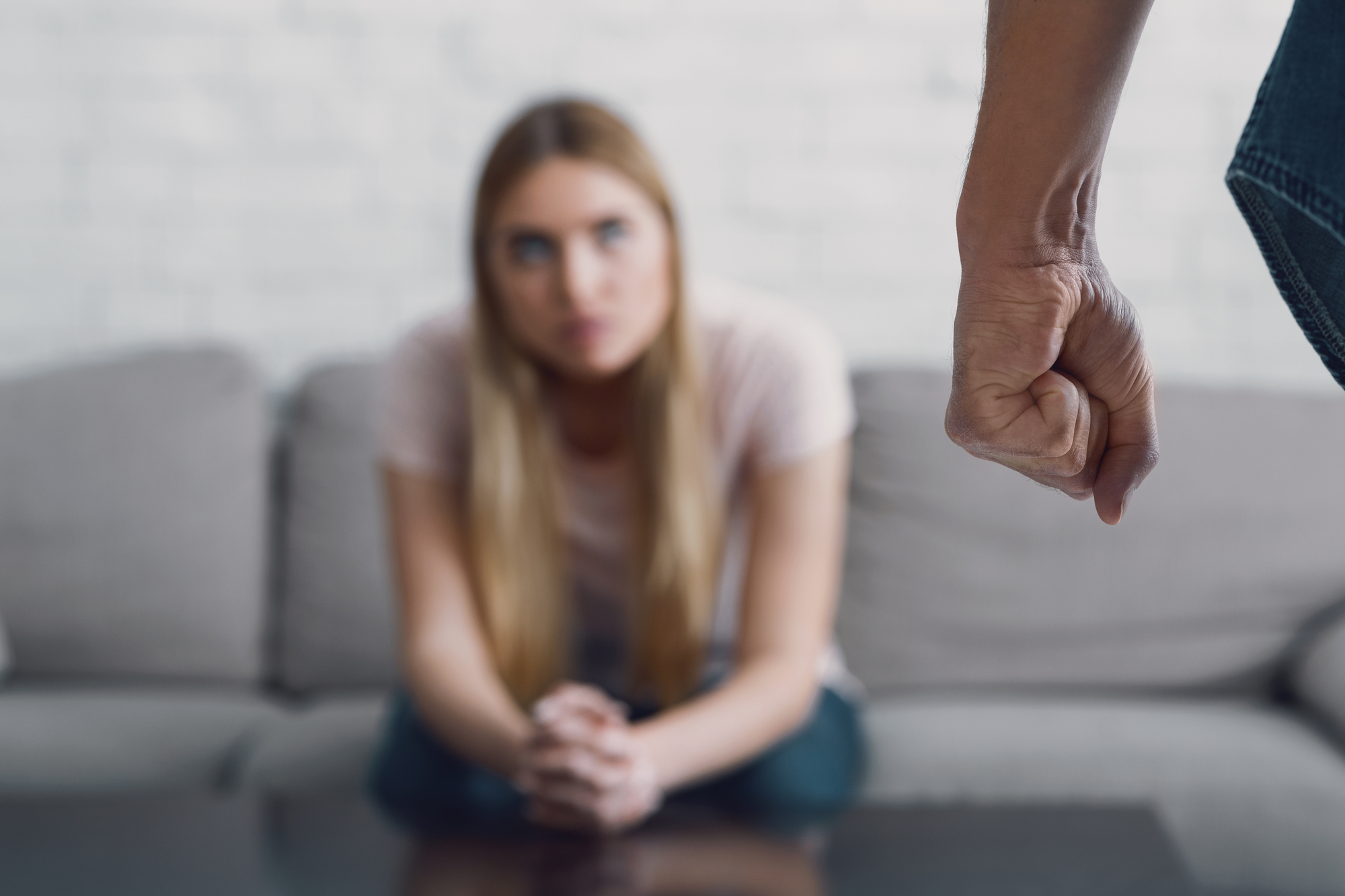 A woman with long blonde hair, sitting on a gray sofa, looks distressed as she focuses on a clenched fist in the foreground. The background features a white brick wall.