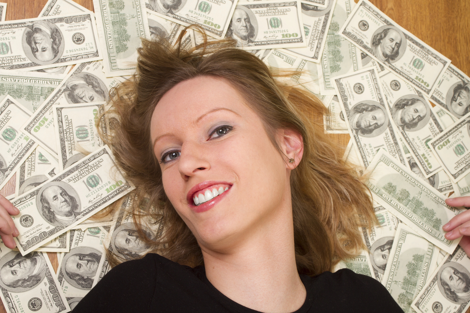 A woman with blonde hair and a black shirt lies on the floor, surrounded by numerous $100 bills. She is smiling and looking at the camera.