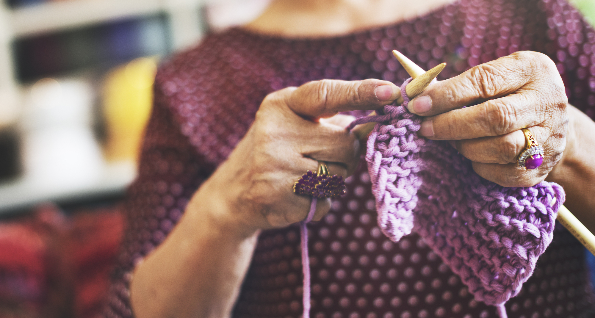 A close-up of an elderly person knitting with wooden needles and purple yarn. The person, wearing a maroon top with white polka dots, is focused on their craft, showcasing the intricate patterns they are creating.