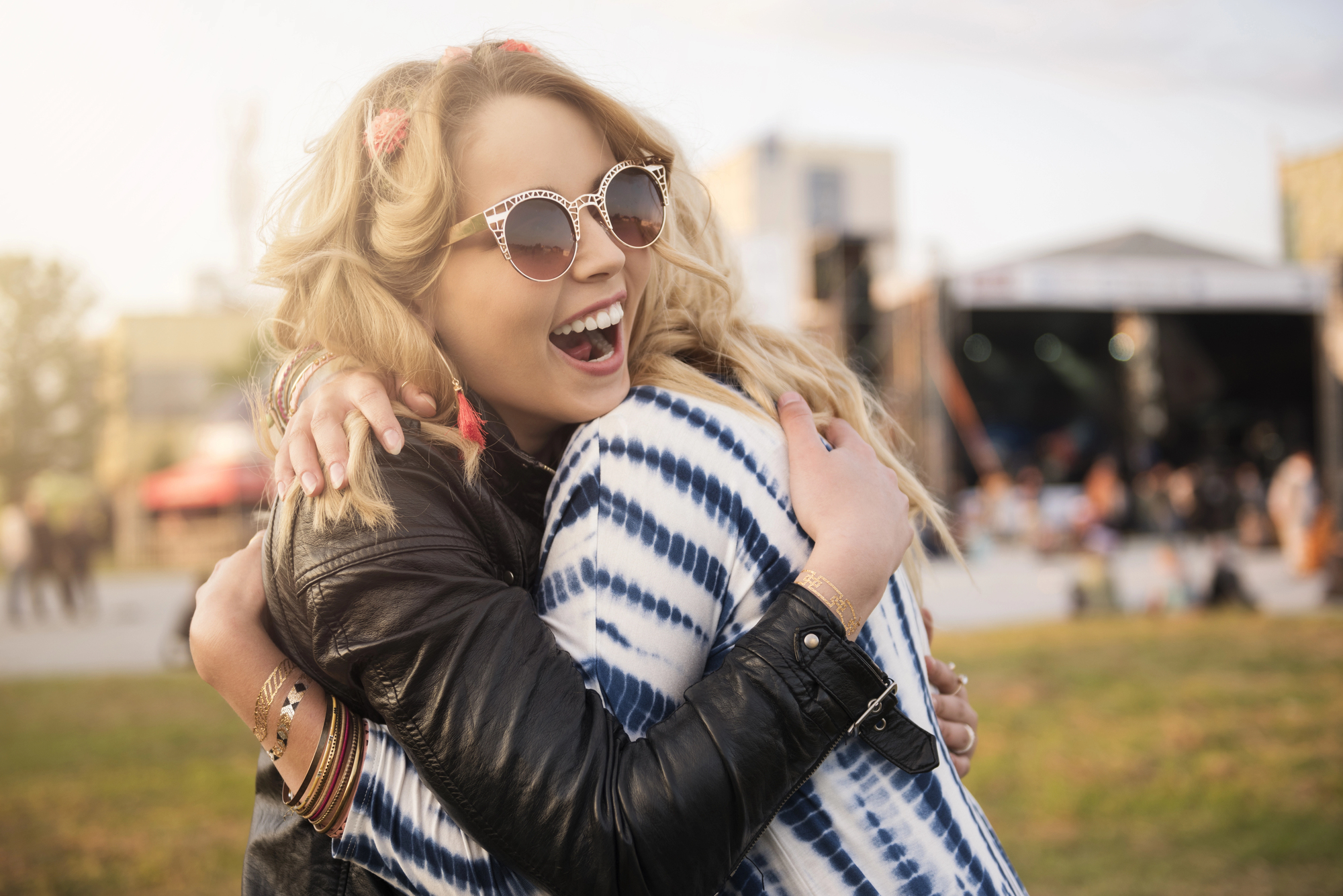 Two women are sharing a joyful hug at an outdoor event. One woman with blonde hair, wearing sunglasses and a leather jacket, has a big smile. The other woman, wearing a blue and white patterned top, has her back to the camera. A stage and crowd are visible in the background.