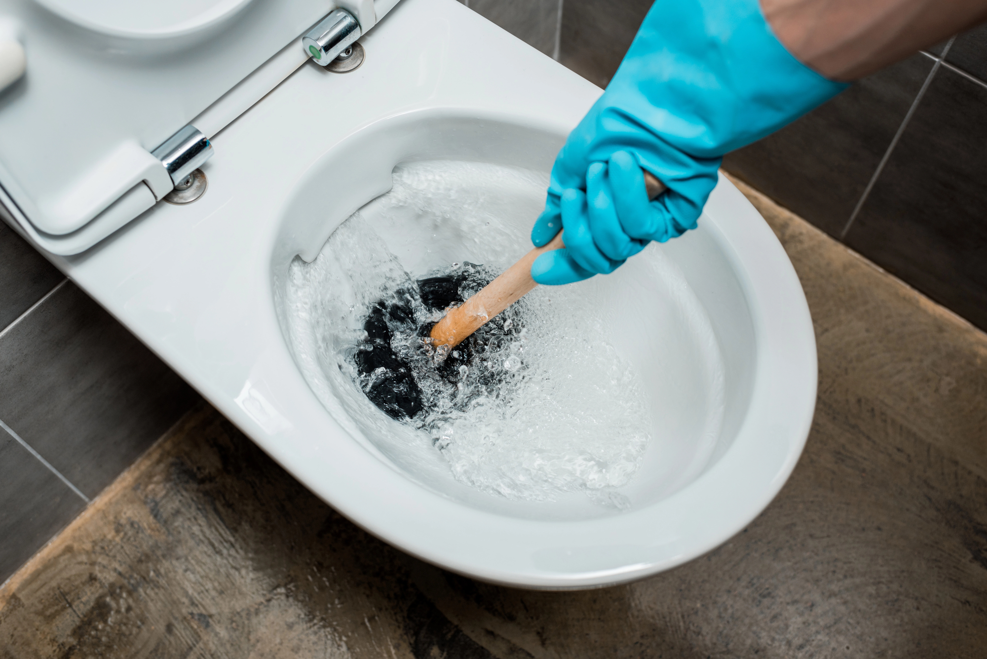 A person wearing blue gloves is using a wooden-handled plunger to unclog a white toilet. Water is swirling in the bowl as they work. The bathroom floor is covered with large, brown tiles. The toilet seat lid is open and visible.