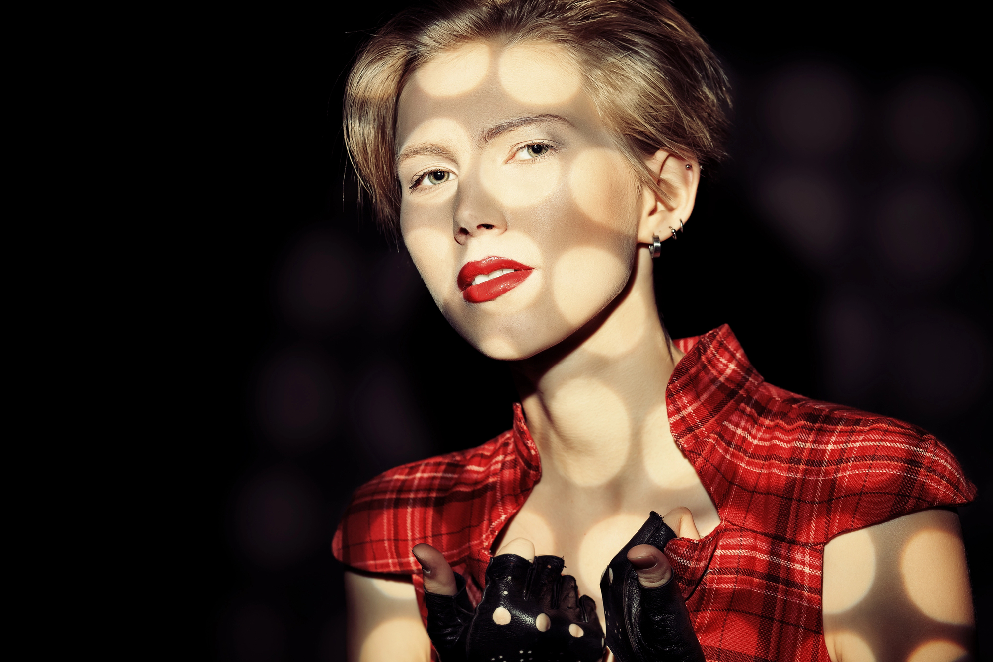 A person with short blonde hair and red lipstick is wearing a sleeveless red plaid shirt and black gloves. They are posing in a way where dappled shadows fall across their face and body, giving an artistic effect against a dark background.