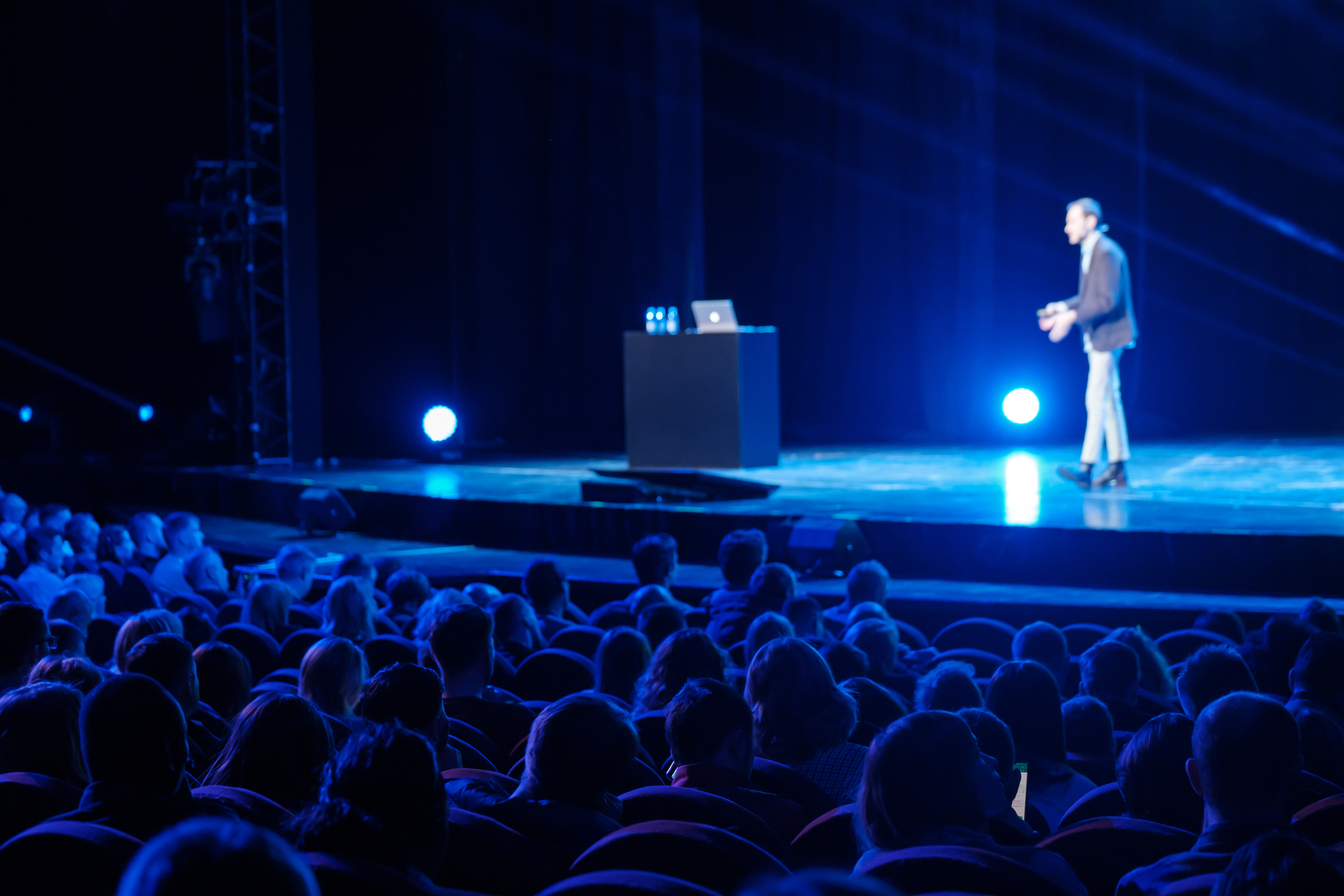 A speaker in business attire addresses a large audience from a stage lit with blue lights. The audience, seated in rows of red chairs, listens attentively. A podium with a laptop and water bottles is placed at the center of the stage.