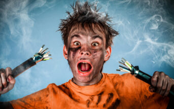 A person with messy hair and a dirty face, wearing a tattered orange shirt, with an expression of shock or surprise. They are holding two electrical cables with exposed wires in front of a smoky background.