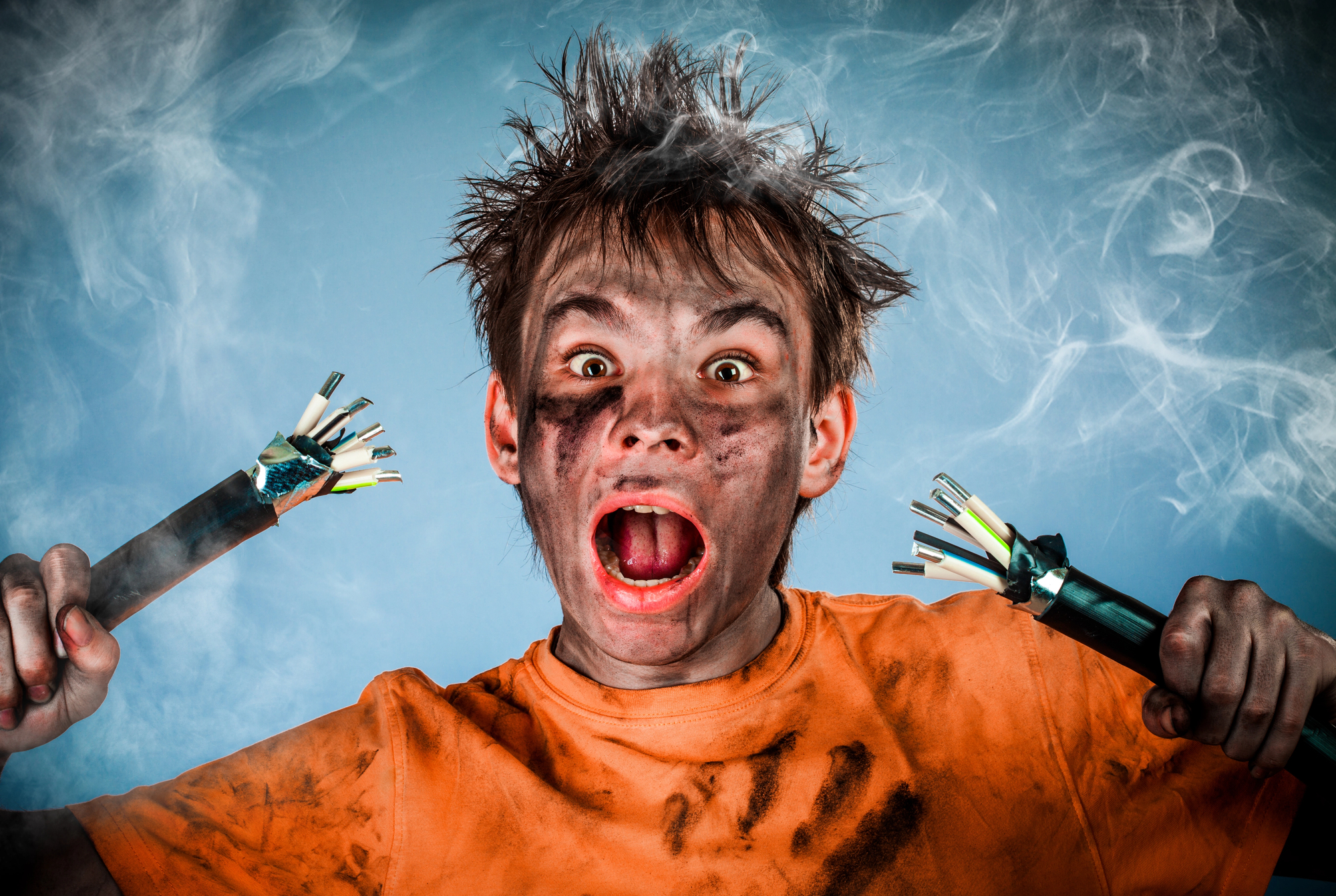 A person with messy hair and a dirty face, wearing a tattered orange shirt, with an expression of shock or surprise. They are holding two electrical cables with exposed wires in front of a smoky background.