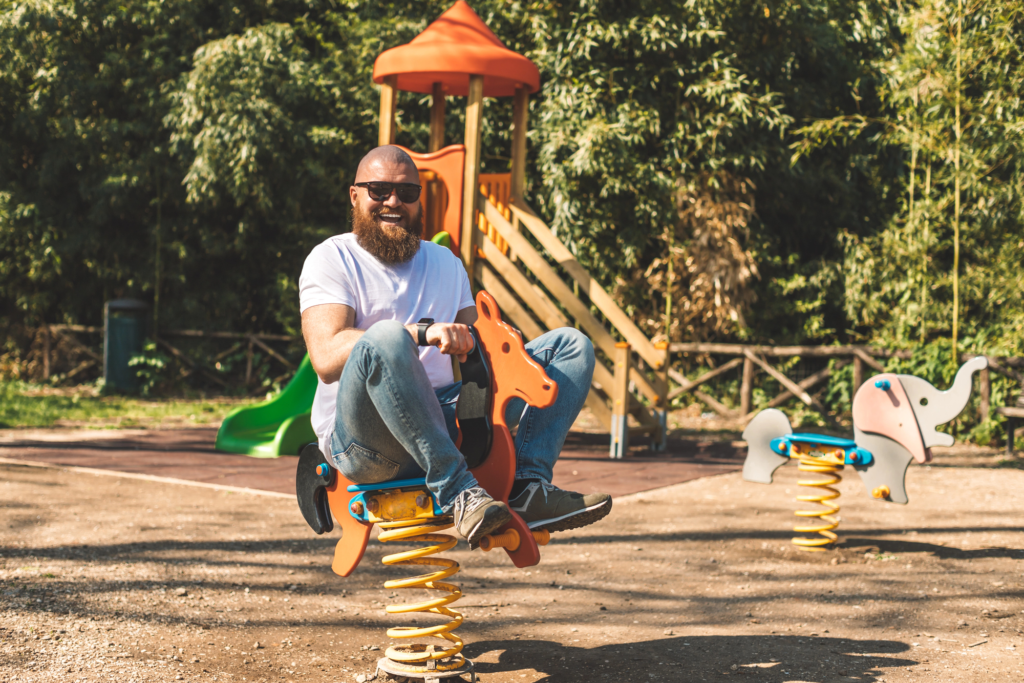 A bearded man with sunglasses, wearing a white t-shirt and jeans, sits joyfully on a spring rocker shaped like a horse in a sunny playground. In the background, there's a slide and climbing structure with green trees surrounding the area.