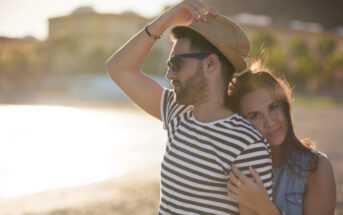 A couple embraces on a sunlit beach. The man, wearing a striped shirt, sunglasses, and a hat, smiles while holding his hat. The woman stands behind him, resting her head on his shoulder with her arms around him, smiling softly. The beach and buildings are blurred in the background.