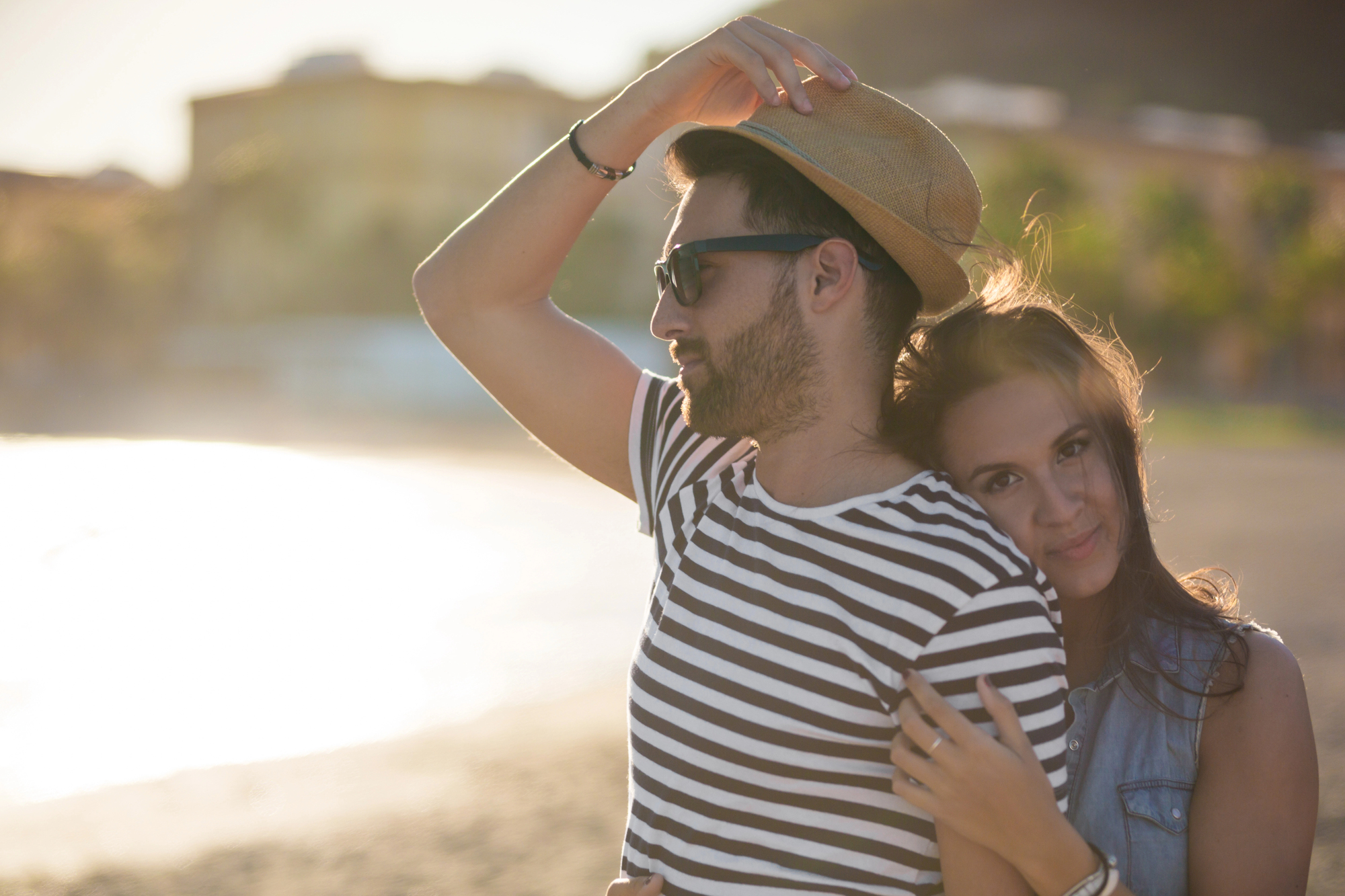 A couple embraces on a sunlit beach. The man, wearing a striped shirt, sunglasses, and a hat, smiles while holding his hat. The woman stands behind him, resting her head on his shoulder with her arms around him, smiling softly. The beach and buildings are blurred in the background.