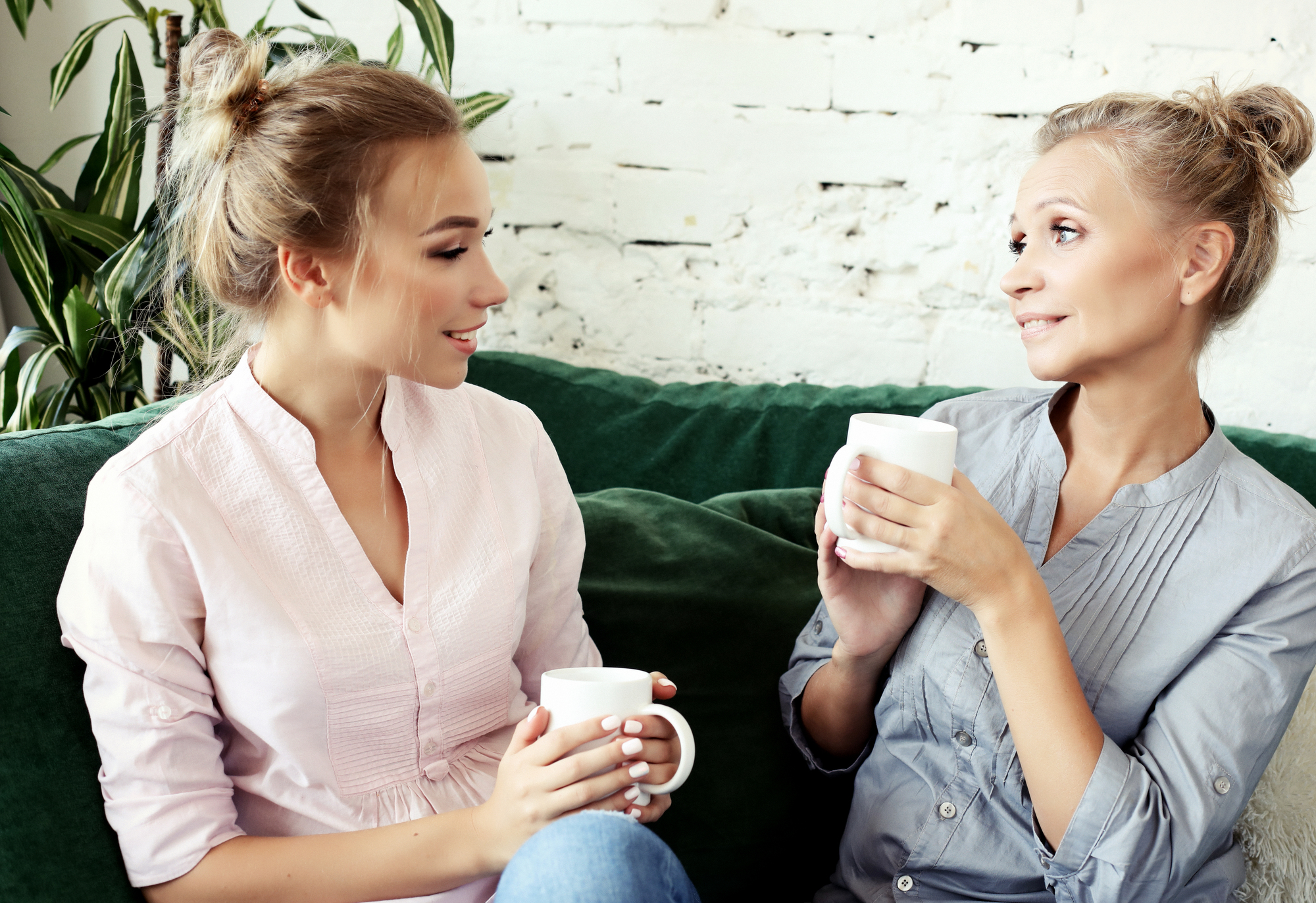 Two women sitting on a green couch, smiling, and holding white mugs. They are engaged in conversation. The room has a white brick wall and green plants. Both women have their hair tied back in bun styles.