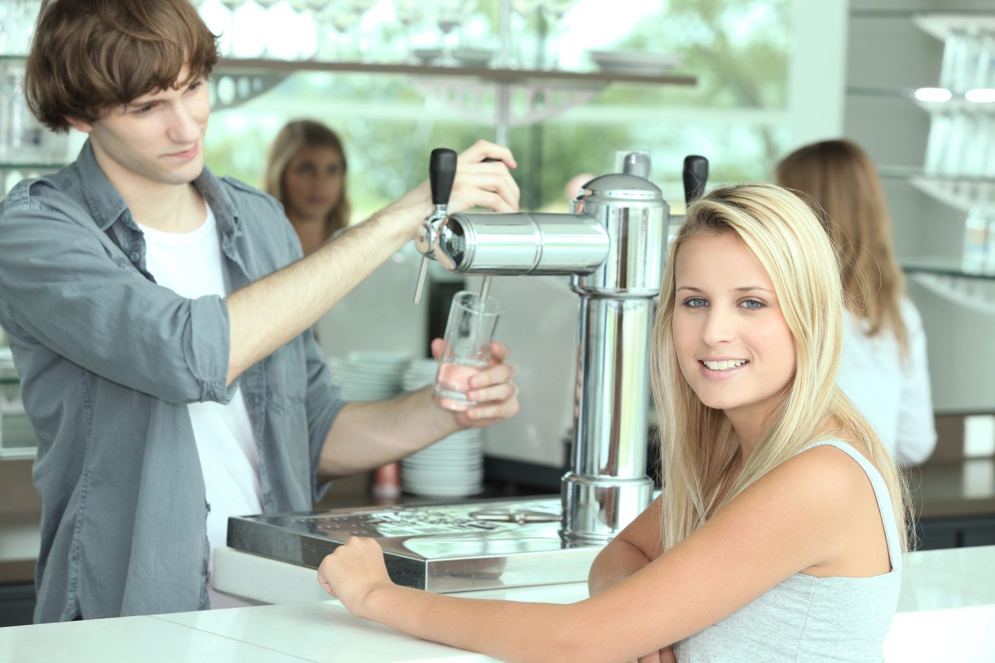 A young man is pouring a drink from a tap while a young woman with long blonde hair smiles at the camera. They are in a bright, modern café with glassware displayed in the background. Another person is visible in the background, slightly out of focus.