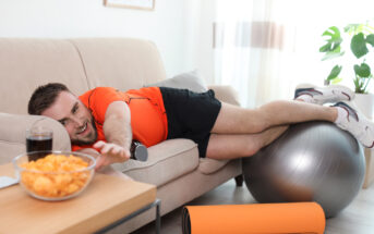 A man in workout attire lies on a couch with his legs on a fitness ball and reaches for a bowl of chips on a nearby table. A glass of soda is next to the bowl. An exercise mat is rolled up on the floor. The scene is set in a bright living room with plants.