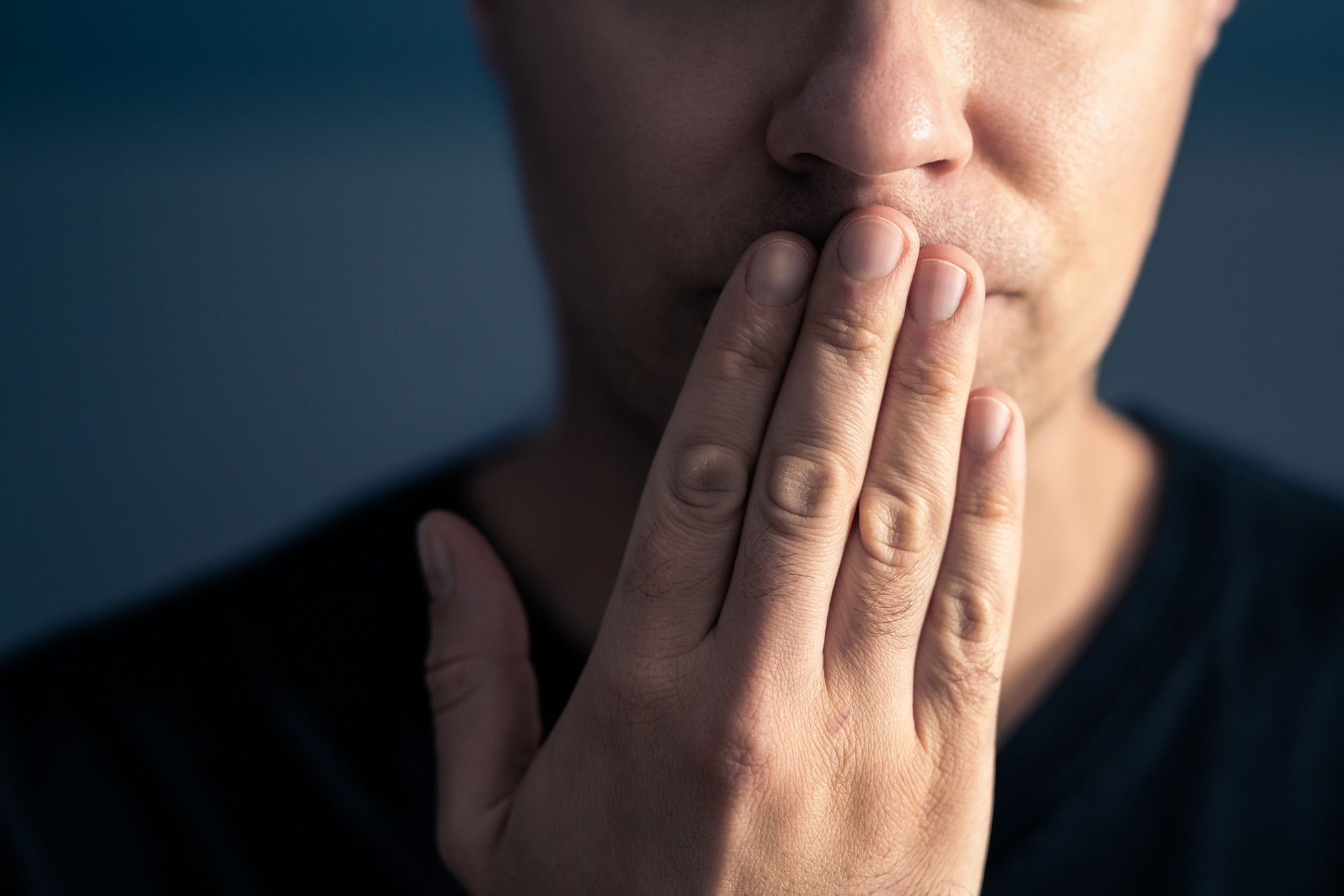 A close-up of a person partially covering their mouth with their hand, suggesting a gesture of silence or contemplation. The person is wearing a dark shirt, and the background is blurred, drawing attention to the hand and facial expression.