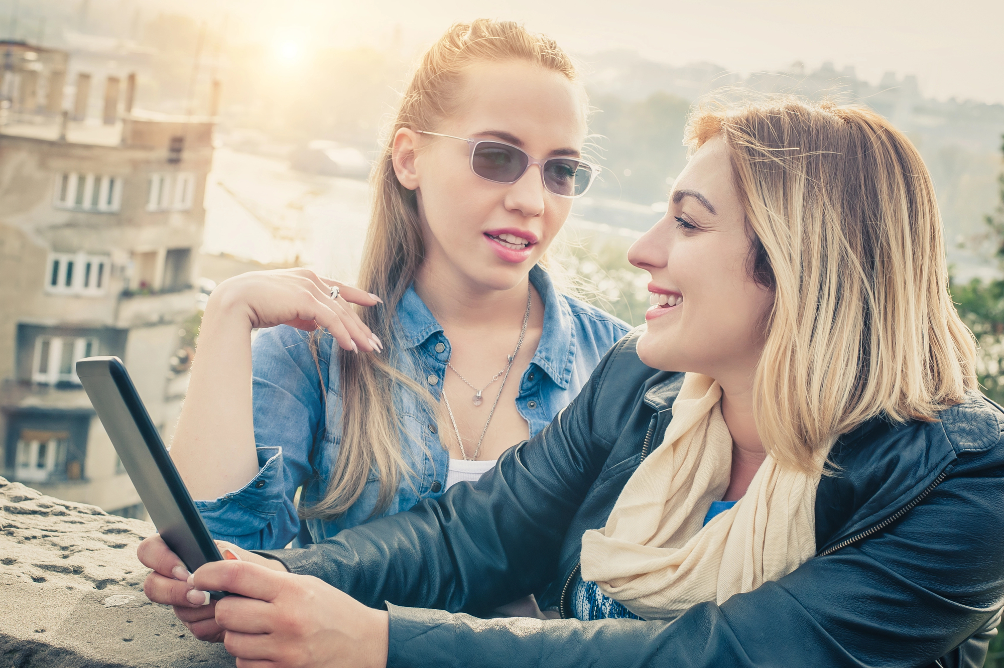 Two women are outdoors, interacting and smiling. One holds a tablet while the other gestures with her hand. Both have light hair, and the background shows buildings and faint sunlight. They appear to be having a pleasant conversation.