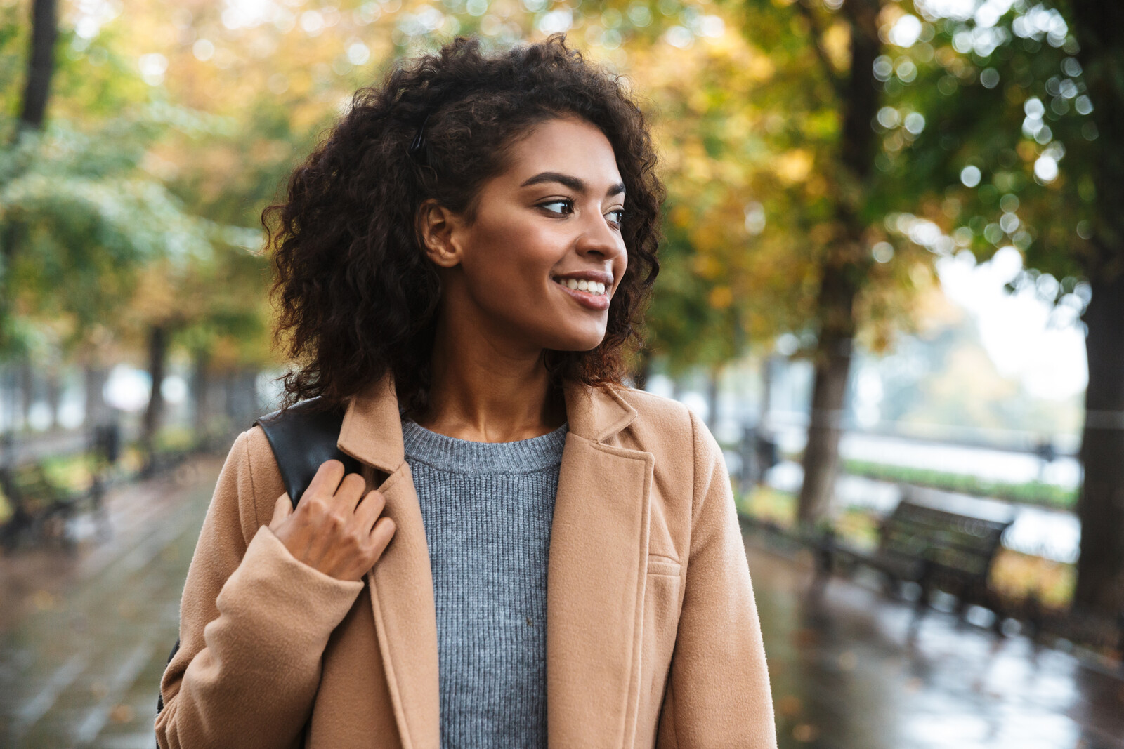 A young woman with curly hair smiles while looking to the side, walking in an outdoor park on a rainy day. She is wearing a tan coat over a gray sweater and holds a strap of a backpack. Trees lining the park path show autumn foliage.