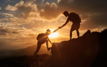 Silhouettes of two hikers at sunset, one reaching down to help the other up a rocky hill. The sky is a mix of orange, yellow, and gray clouds, with the sun near the horizon, creating a dramatic and inspiring scene.