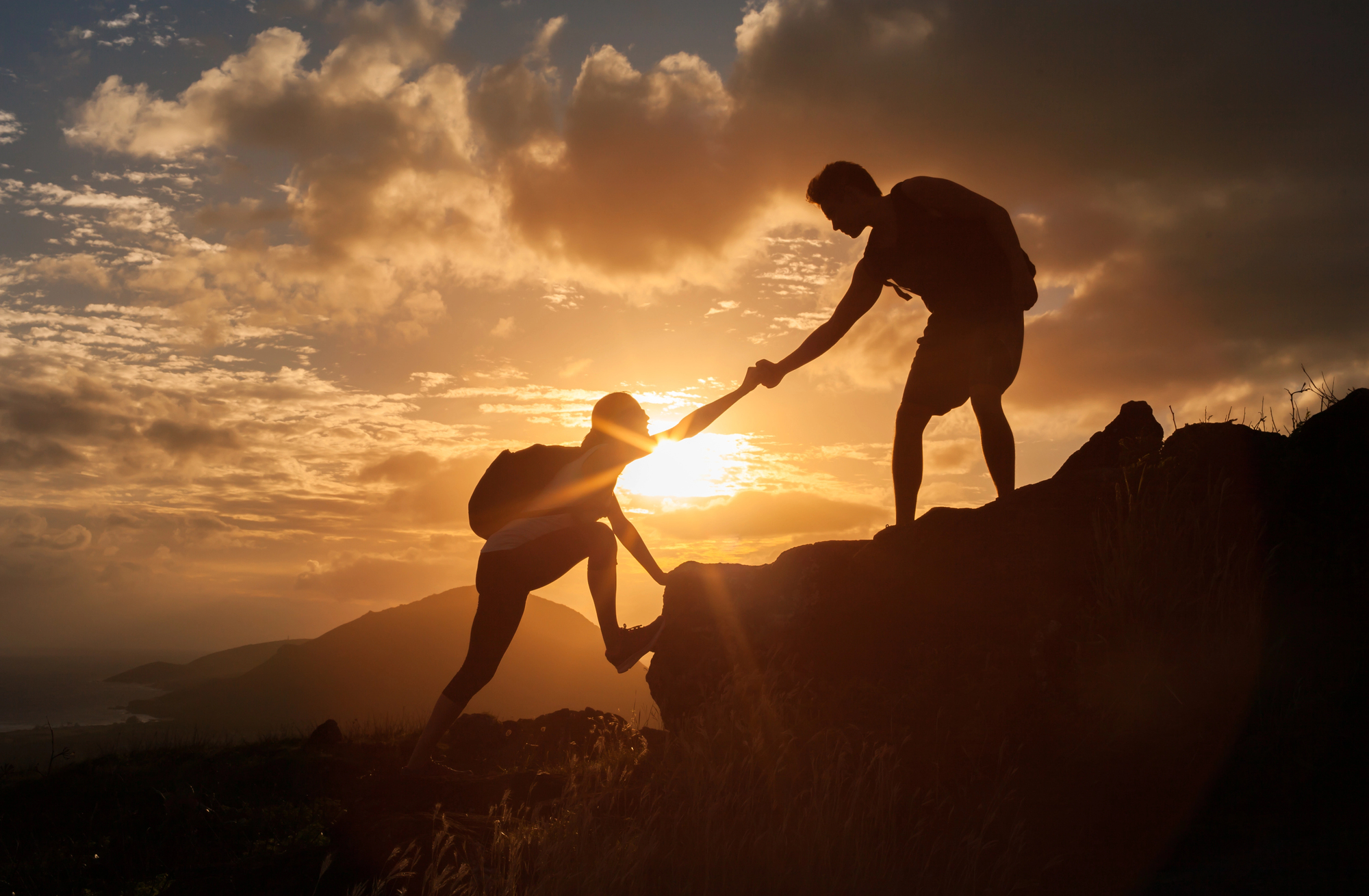 Silhouettes of two hikers at sunset, one reaching down to help the other up a rocky hill. The sky is a mix of orange, yellow, and gray clouds, with the sun near the horizon, creating a dramatic and inspiring scene.