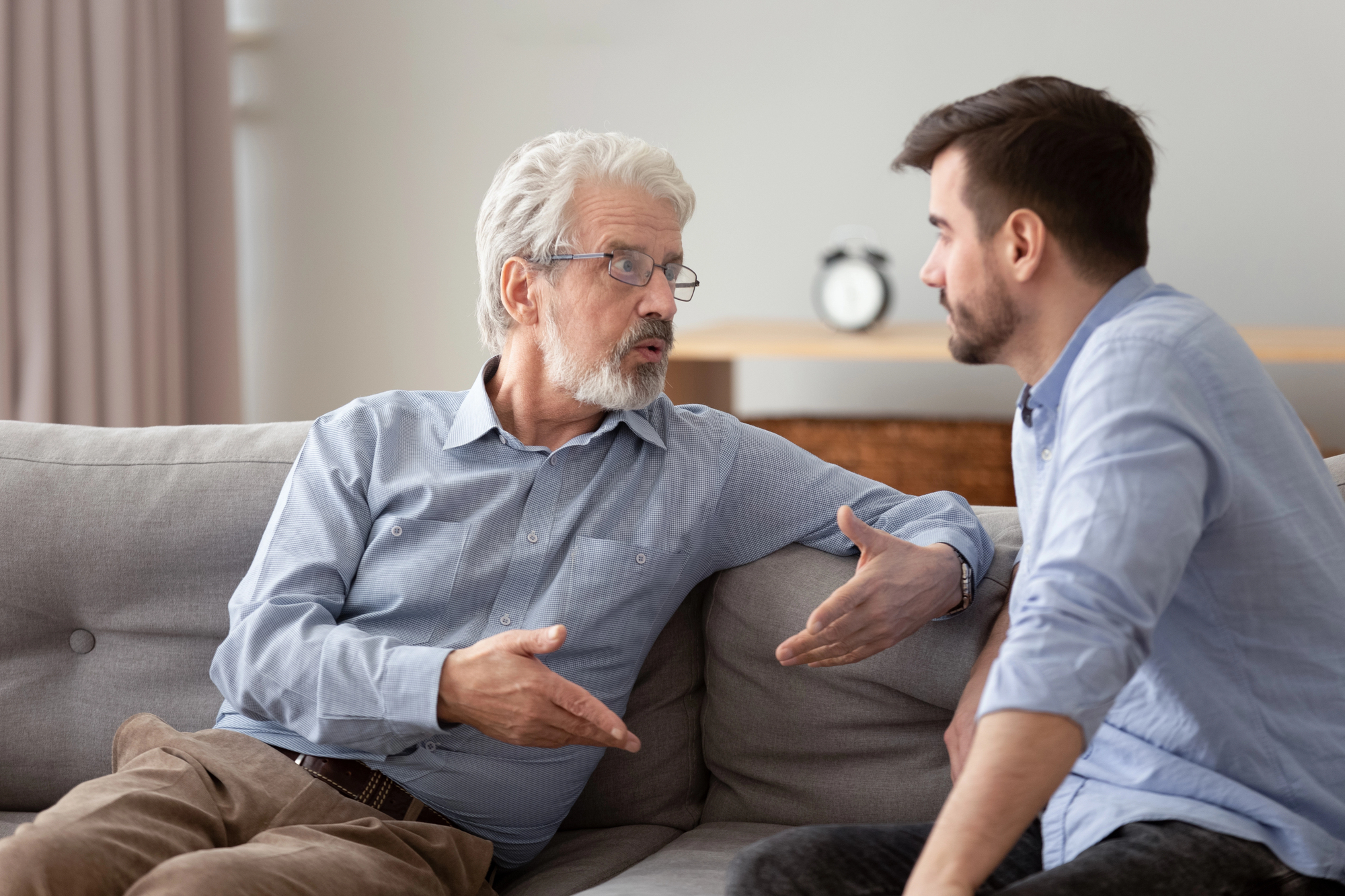 An older man with white hair and glasses is engaged in a serious conversation with a younger man with a beard. They are sitting on a gray couch in a well-lit room, facing each other, with the older man gesturing with his hands.