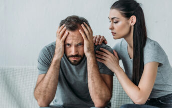 A man sits on a couch with his head in his hands, appearing distressed. A woman sits beside him, gently placing her hand on his shoulder, offering comfort and support. Both are dressed in casual gray attire. The background is simple and uncluttered.