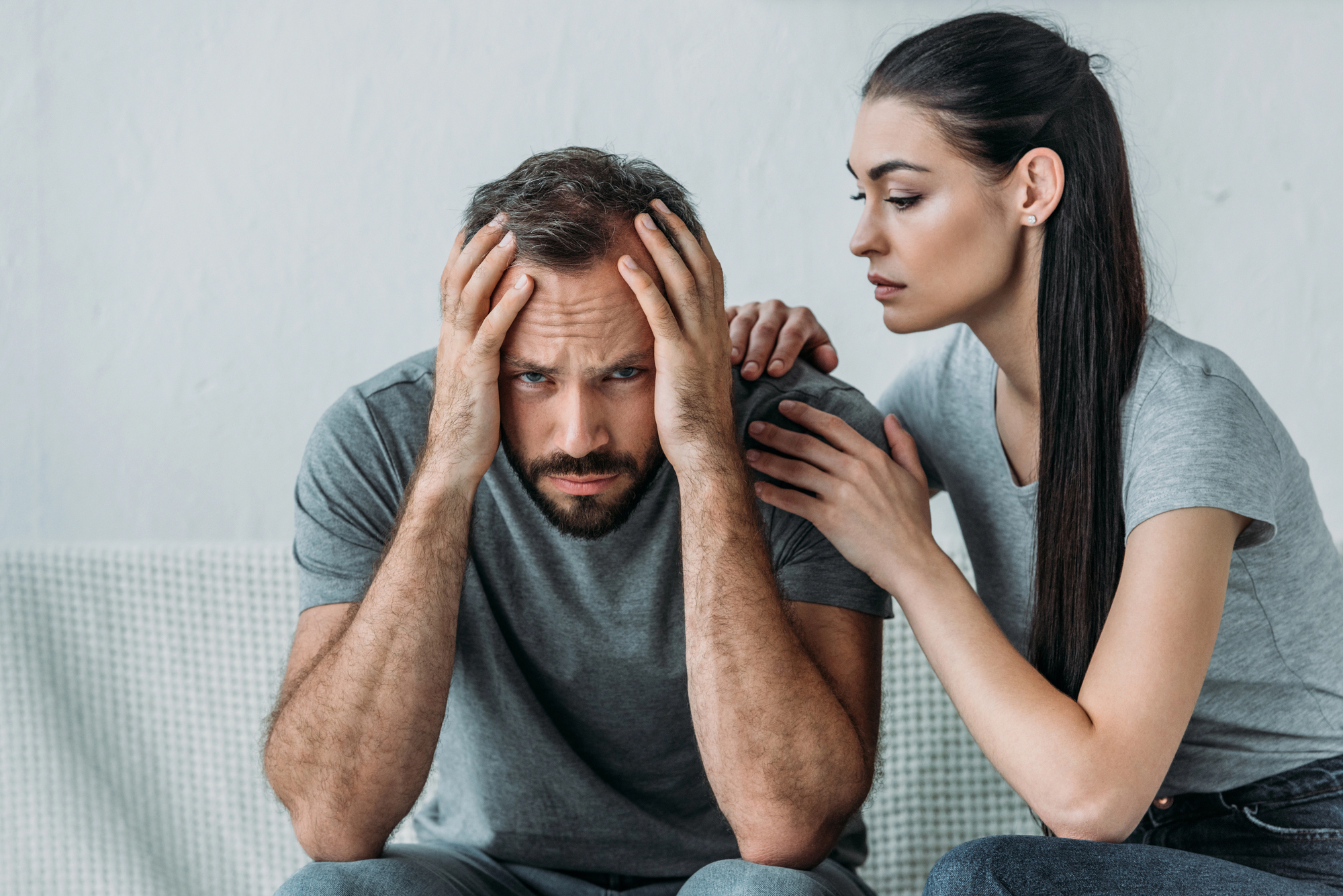 A man sits on a couch with his head in his hands, appearing distressed. A woman sits beside him, gently placing her hand on his shoulder, offering comfort and support. Both are dressed in casual gray attire. The background is simple and uncluttered.