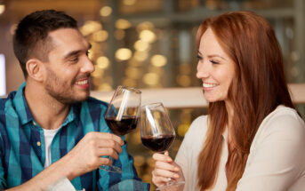 A man and a woman smiling and clinking wine glasses, each filled with red wine. The man has short dark hair and wears a green and blue checkered shirt. The woman has long red hair and wears a white top. They appear to be in a warmly lit setting.