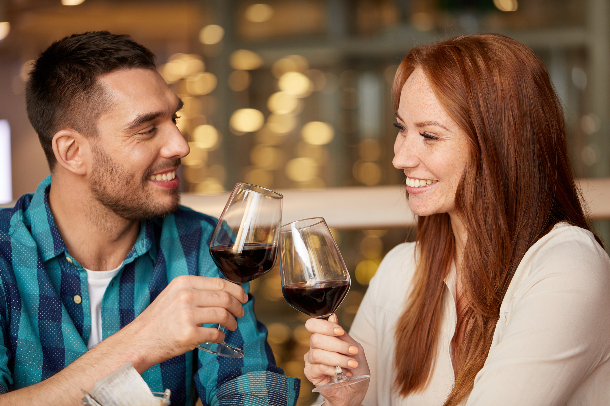 A man and a woman smiling and clinking wine glasses, each filled with red wine. The man has short dark hair and wears a green and blue checkered shirt. The woman has long red hair and wears a white top. They appear to be in a warmly lit setting.