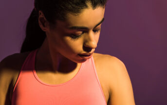 A woman wearing a pink athletic top looks downward with a neutral expression. The background is a soft gradient of purple and pink, creating a serene and focused atmosphere. Her hair is tied back, and the lighting creates gentle shadows on her face and shoulder.